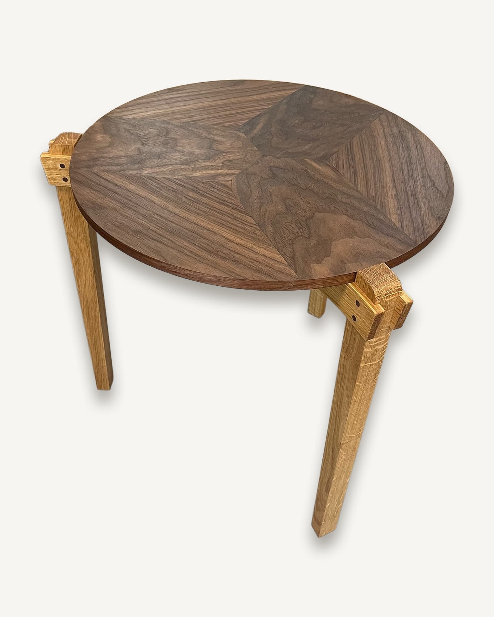 Round walnut top with geometric book-matched veneer pattern, supported by three straight oak legs with visible peg joinery