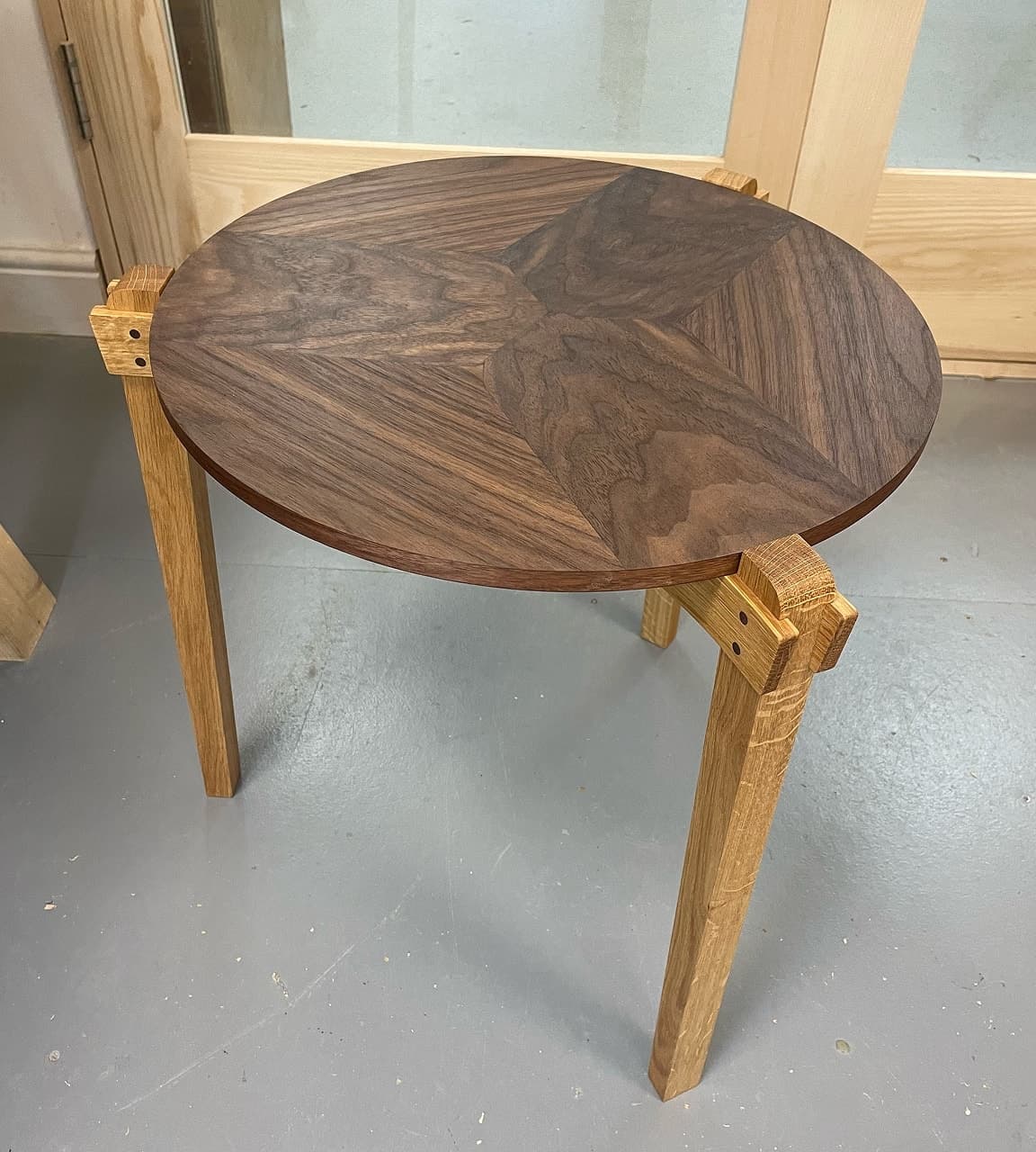 Round walnut table top with book-matched grain on three tapered pale legs, on a workshop floor.