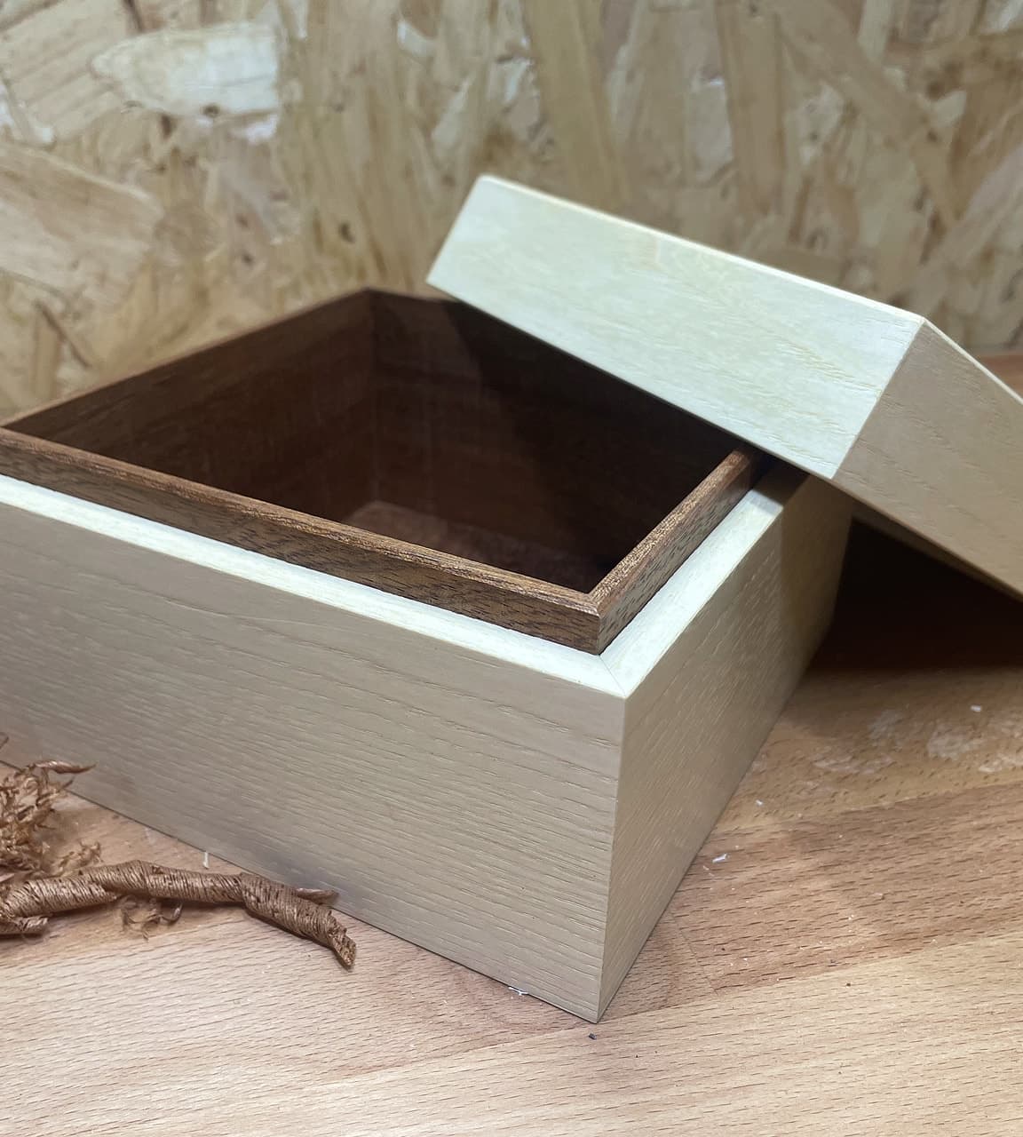Pale oak box with the lid tilted off, showing a dark timber lining, on a bench against a boarded wall.