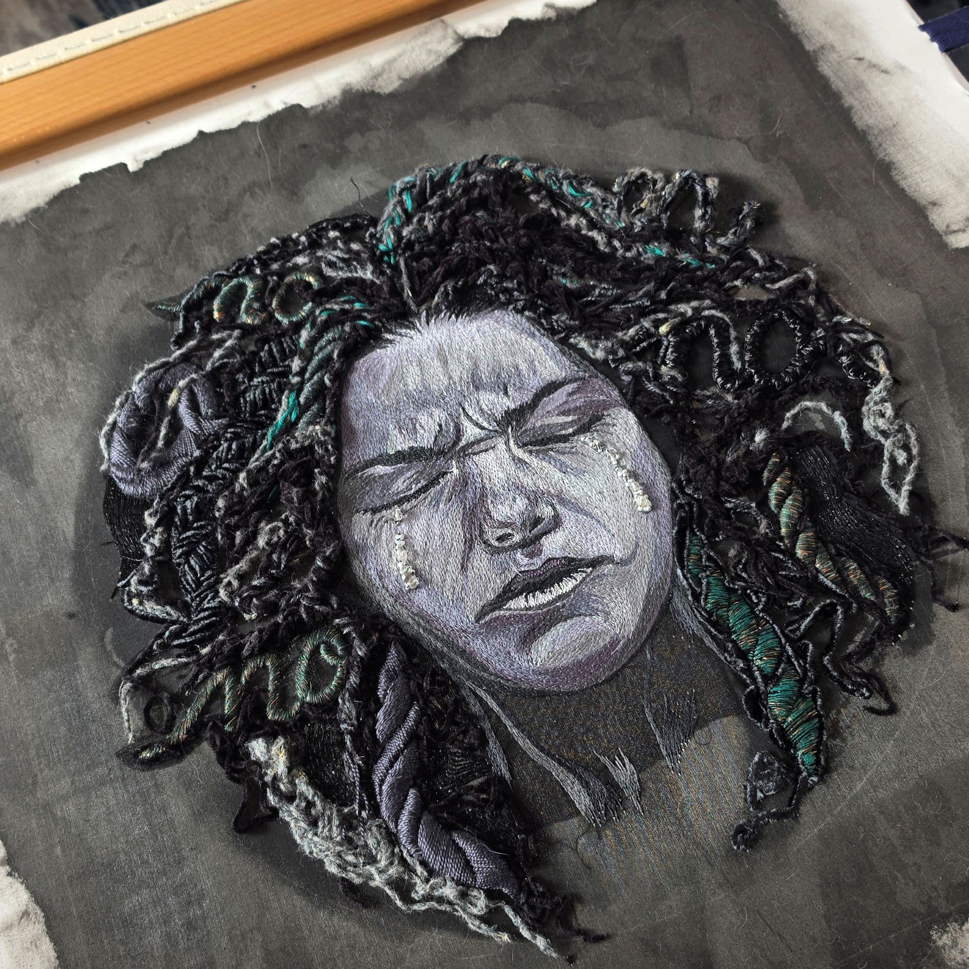 Hand-embroidered face in greyscale thread with raised cord hair, mounted on a grey ground