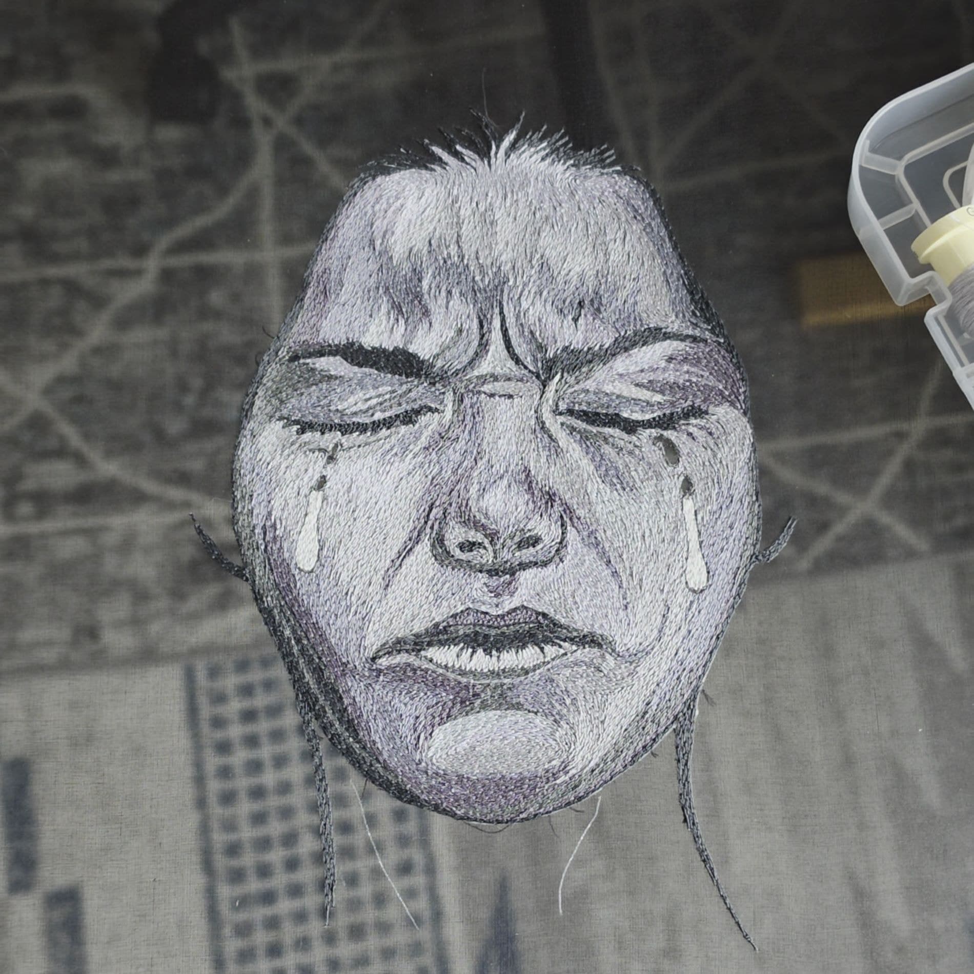 Embroidered face with stitched tears, worked in white thread on a grey woven ground