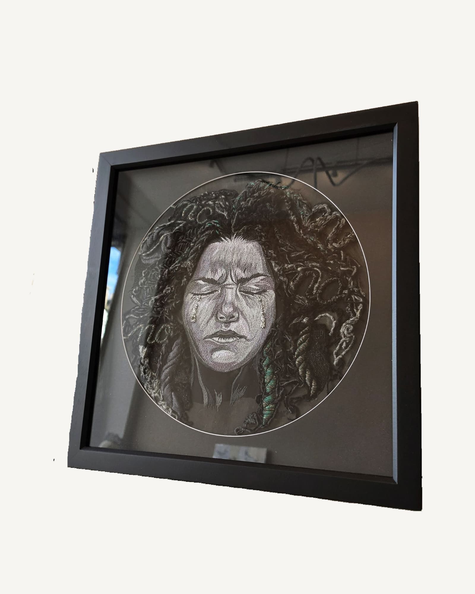 Two framed textile artworks leaning against a wall, each a greyscale embroidered face in a circular composition