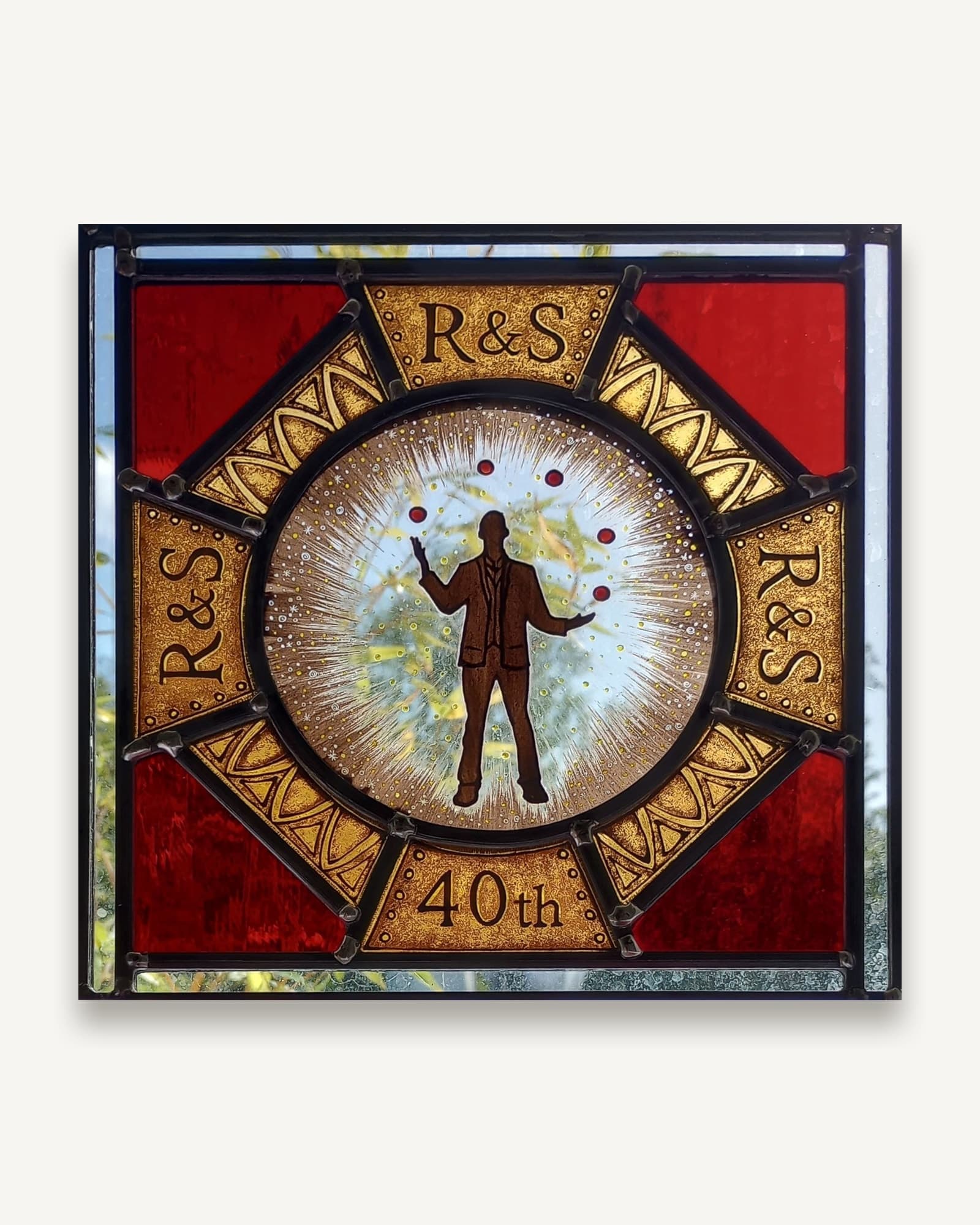 A painted and fired stained glass anniversary panel, a juggling figure ringed by eight amber segments carrying a pair of initials.