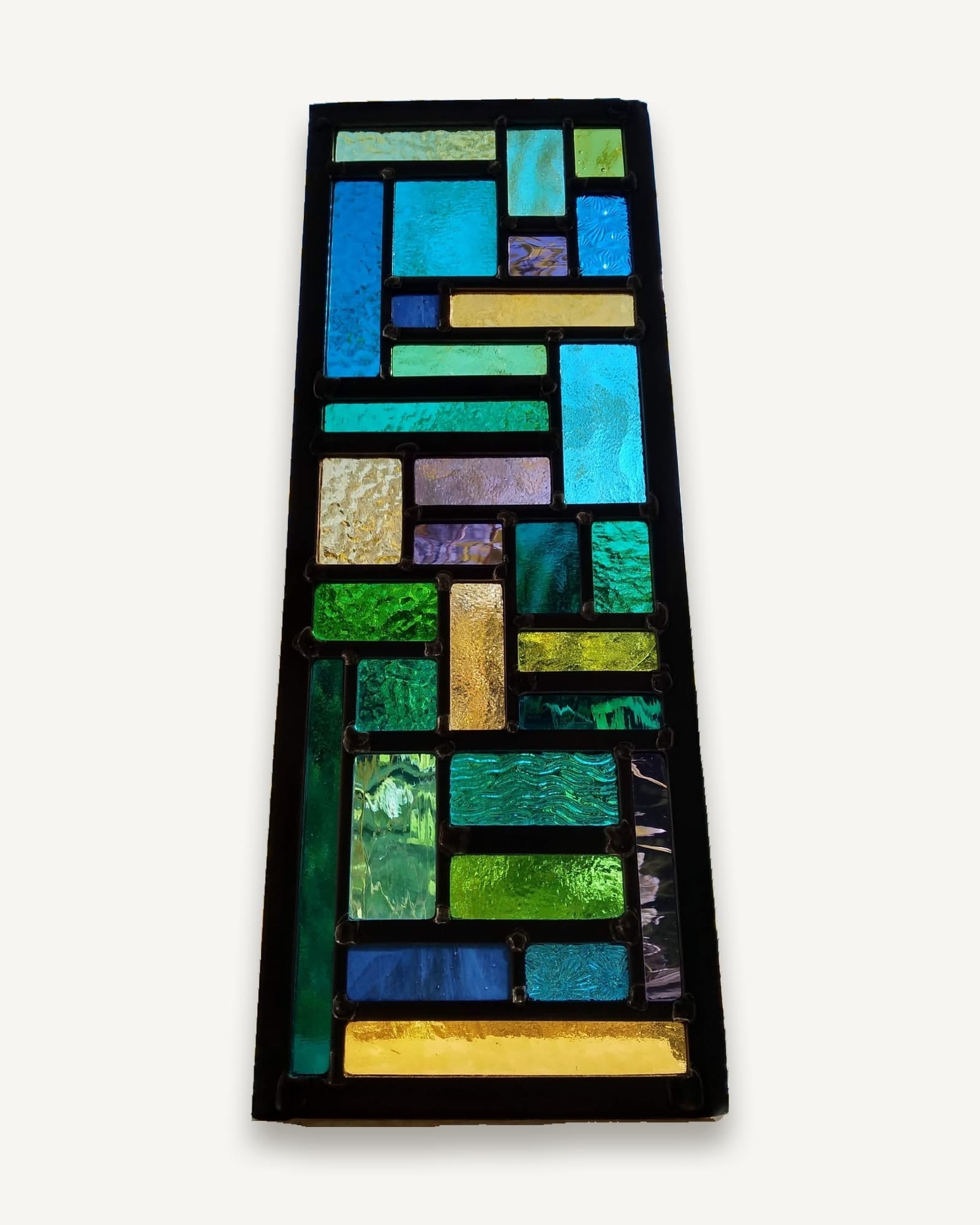 A tall tapering leaded panel of blue, green, violet and amber glass on a wooden base, several pieces textured or iridescent.