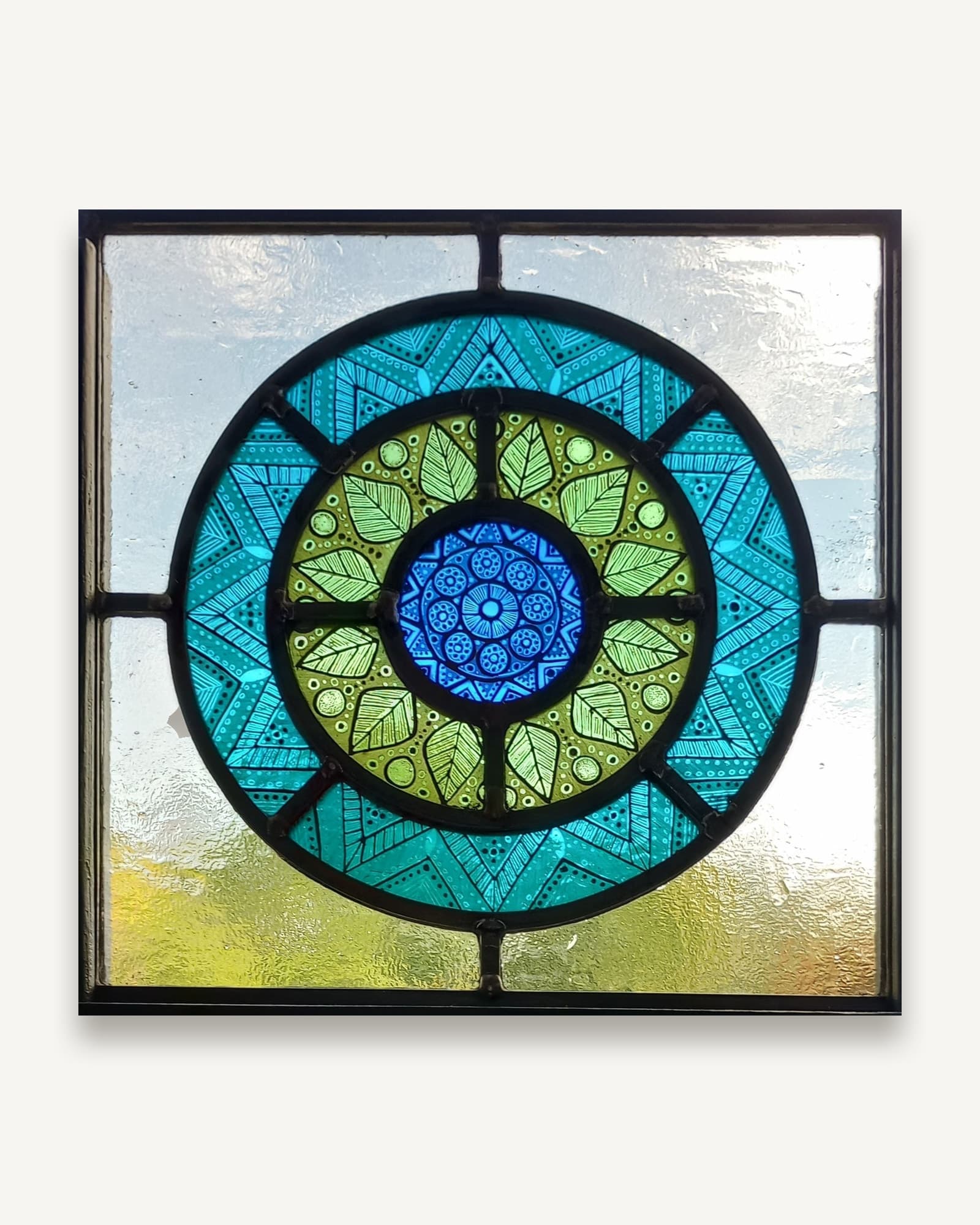A leaded panel with a ring of painted green leaves on teal glass around a blue rosette, set into pale textured glass.