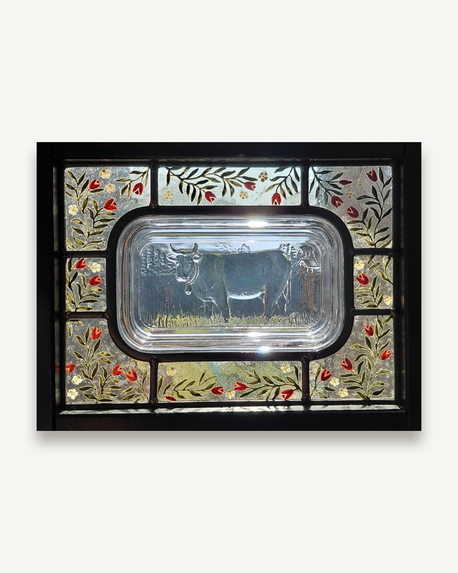 A leaded stained glass panel built around a vintage pressed-glass butter dish lid, its moulded cow framed in painted flowers.