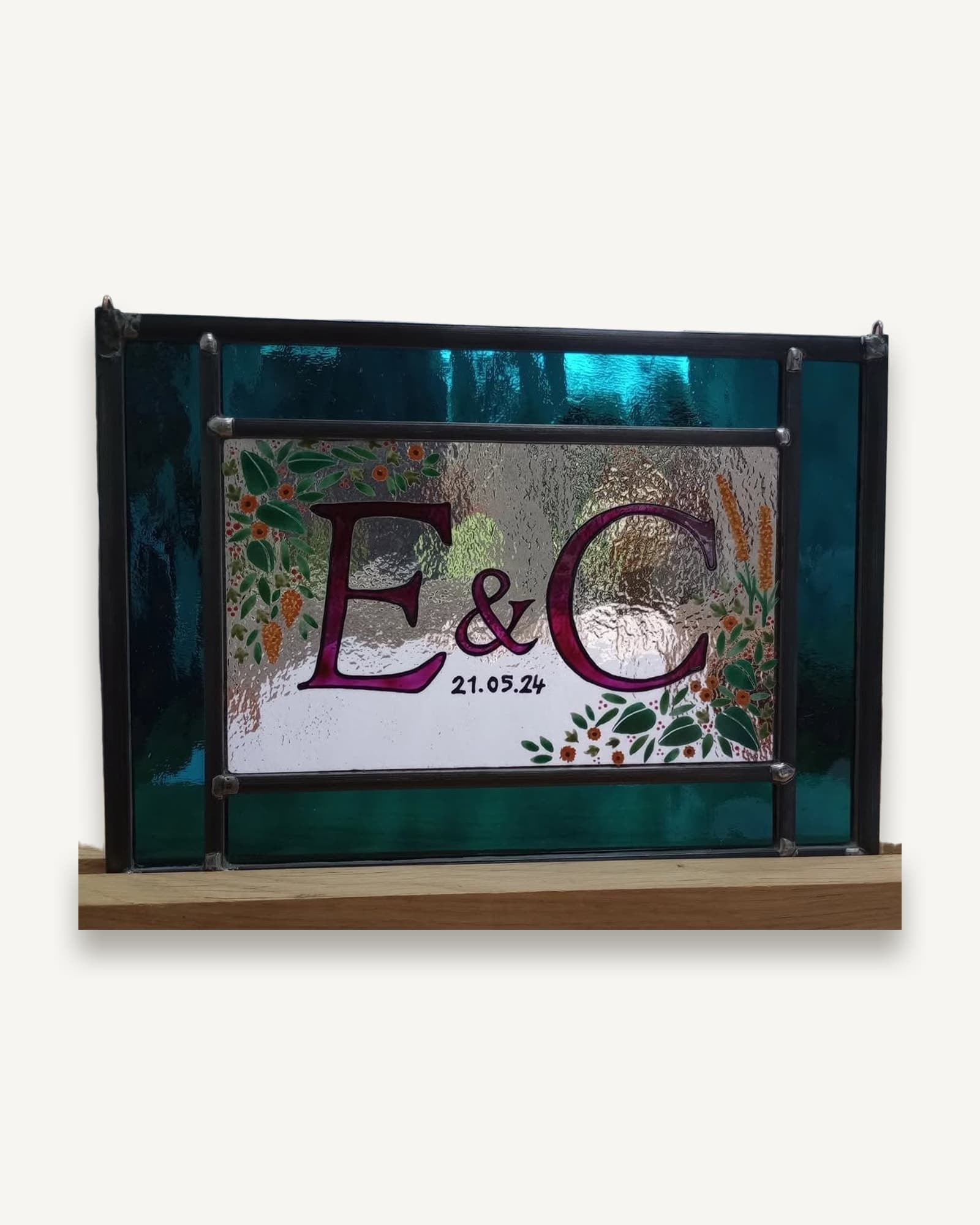 A wedding commission in stained glass: painted initials and the date ringed by flowers, on a teal leaded panel.
