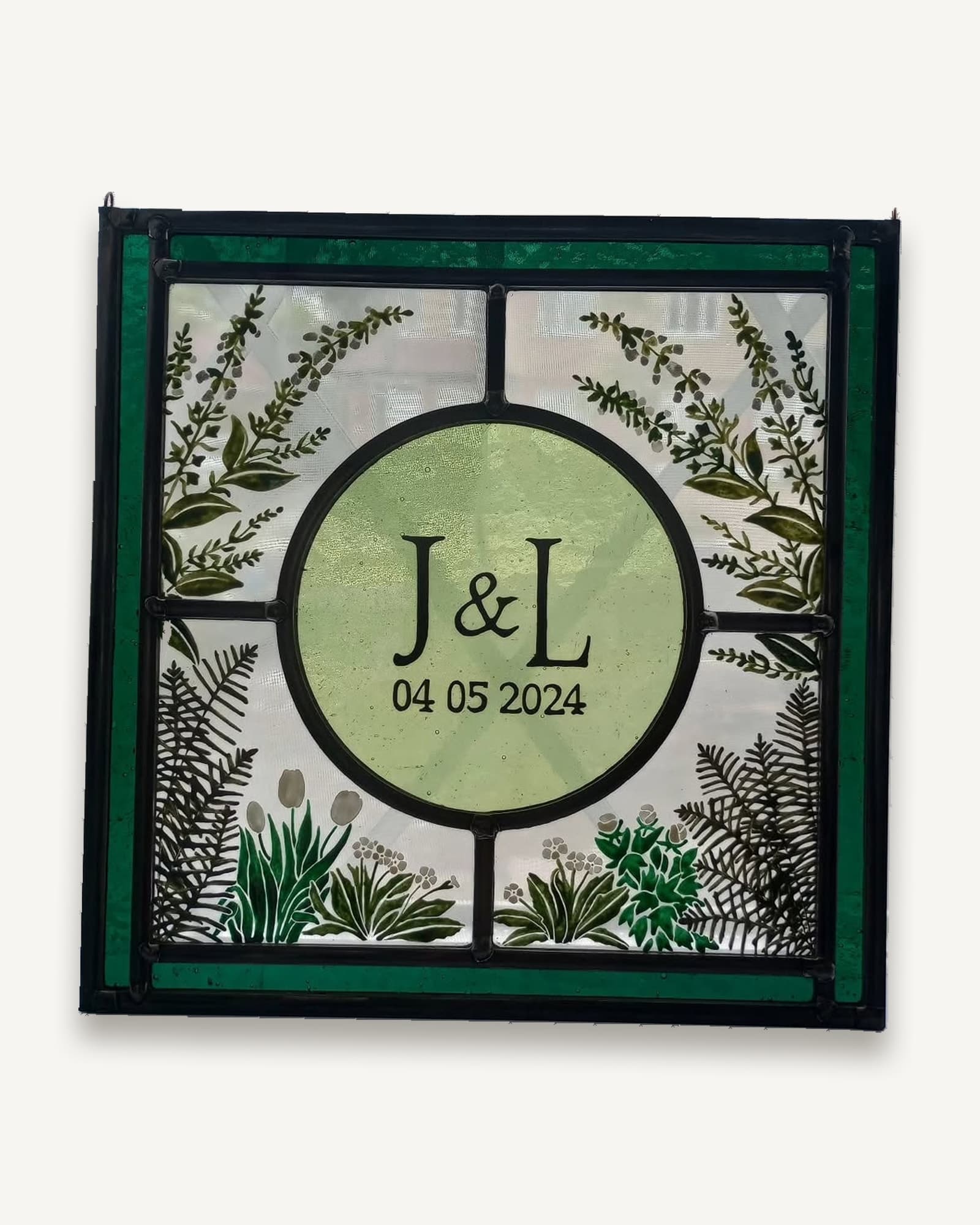 A stained glass wedding gift panel: initials and date in a painted roundel framed by ferns and seed heads.