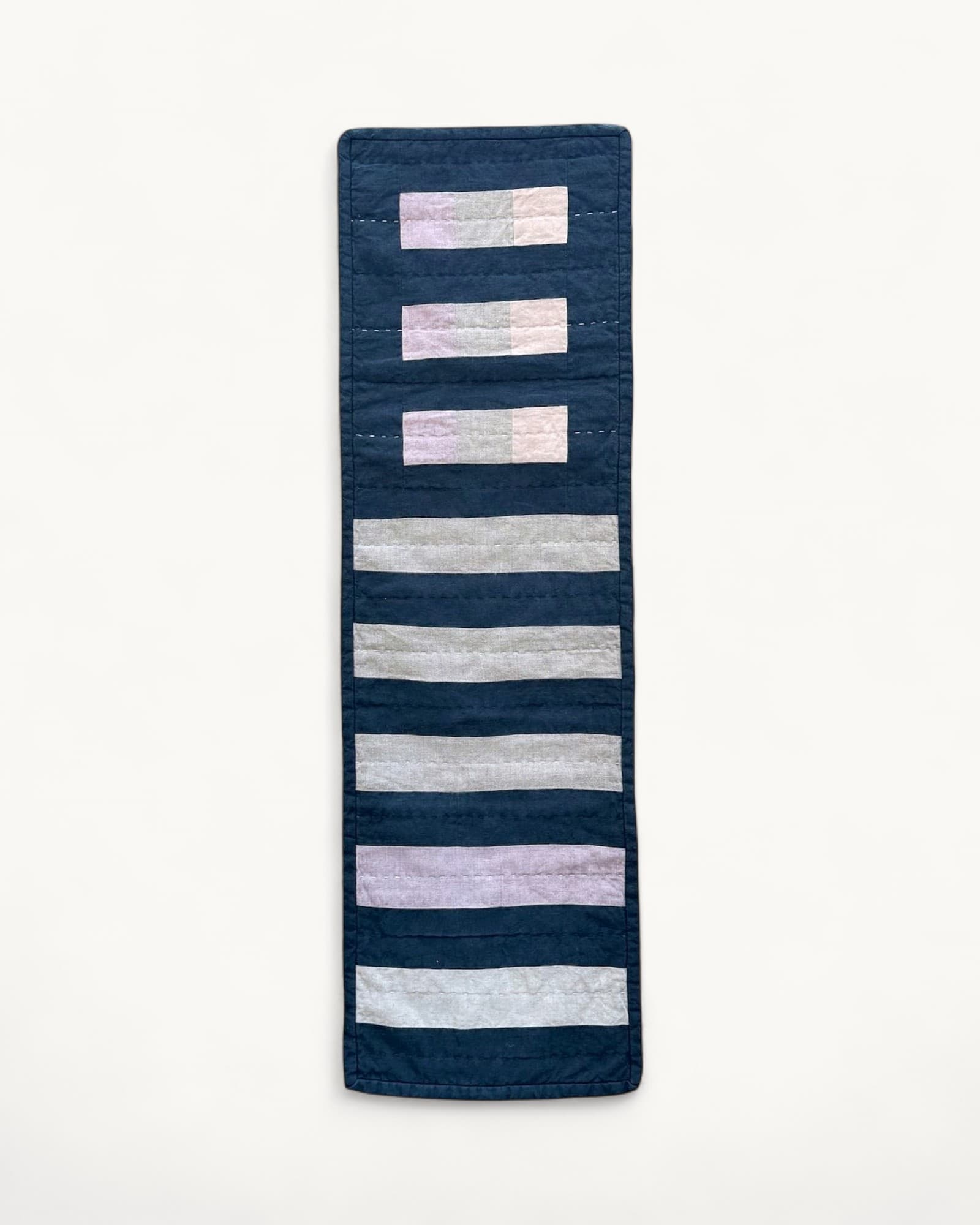 Narrow vertical quilted wall hanging in dark blue linen with horizontal stripes of pale pink, grey and cream fabric,