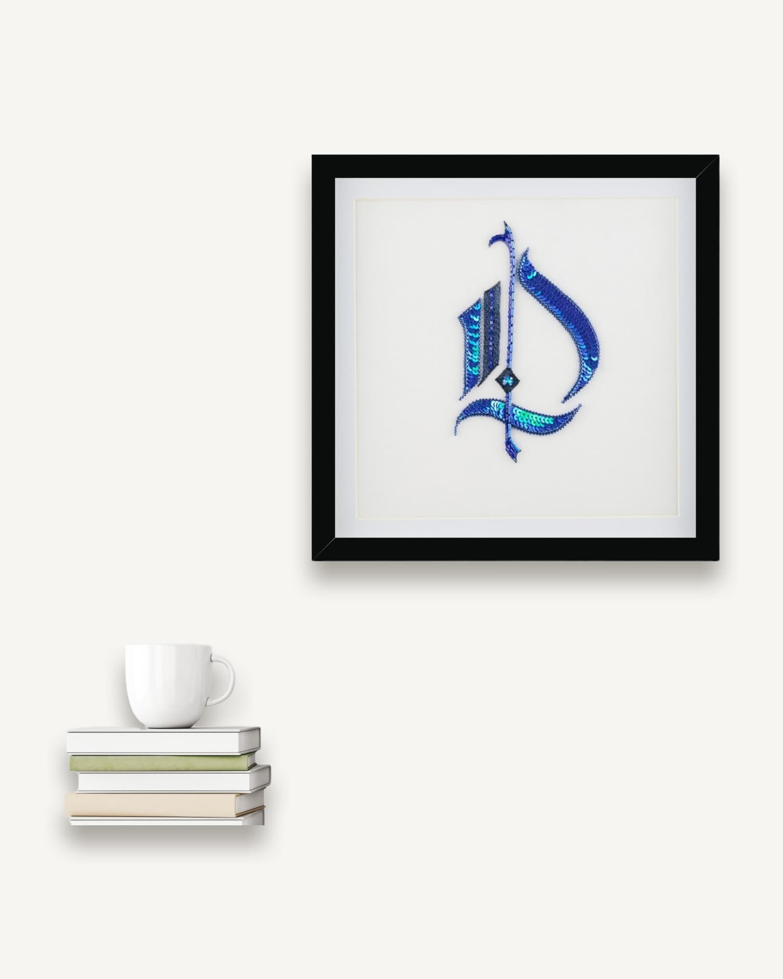 Framed embroidery in black frame with white mount showing stylized Gothic letter in blue and turquoise thread with textured