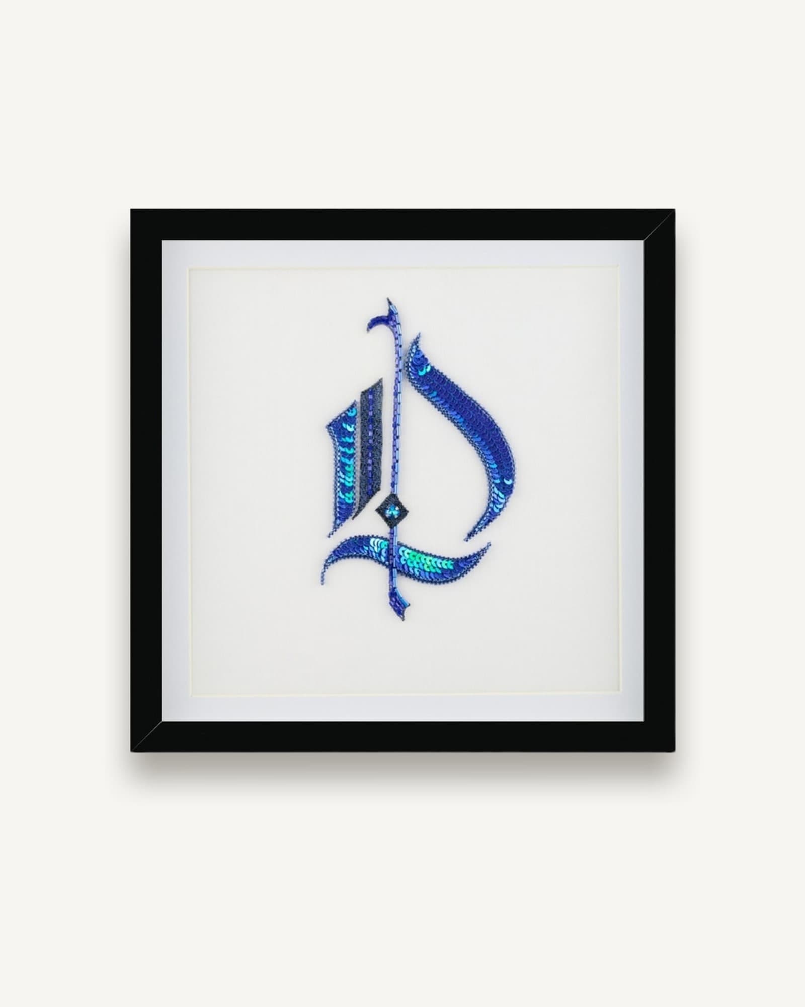 Framed embroidery in black frame with white mount showing stylized Gothic letter in blue and turquoise thread with textured