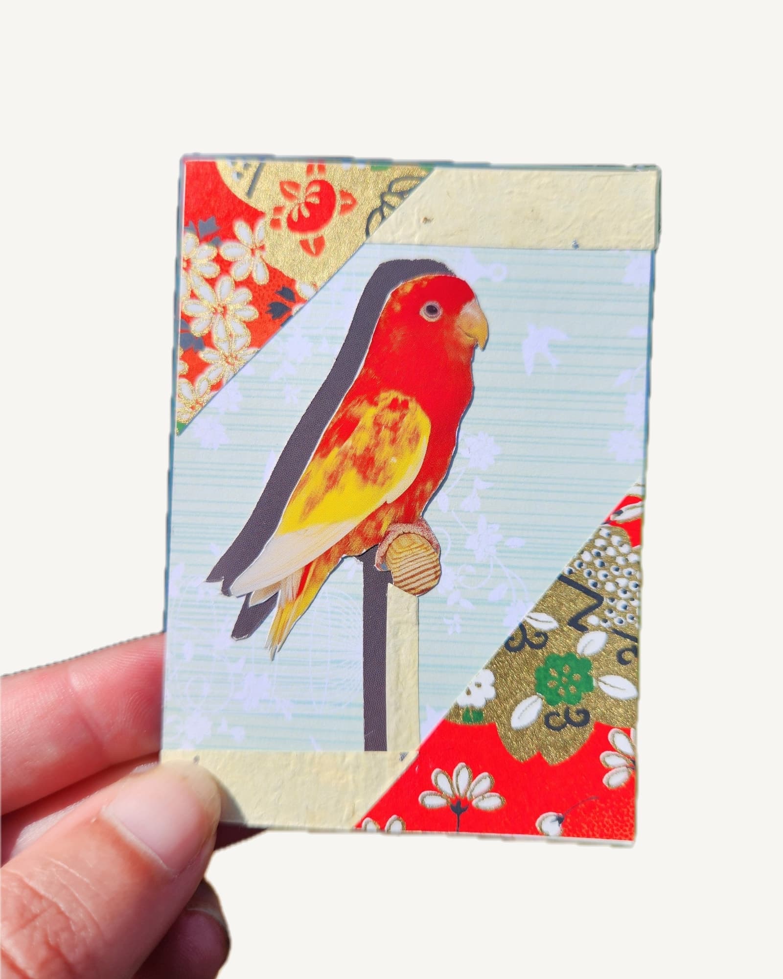 A red and yellow lovebird cut by hand from a secondhand bird book, set against mint green card and red floral origami paper.