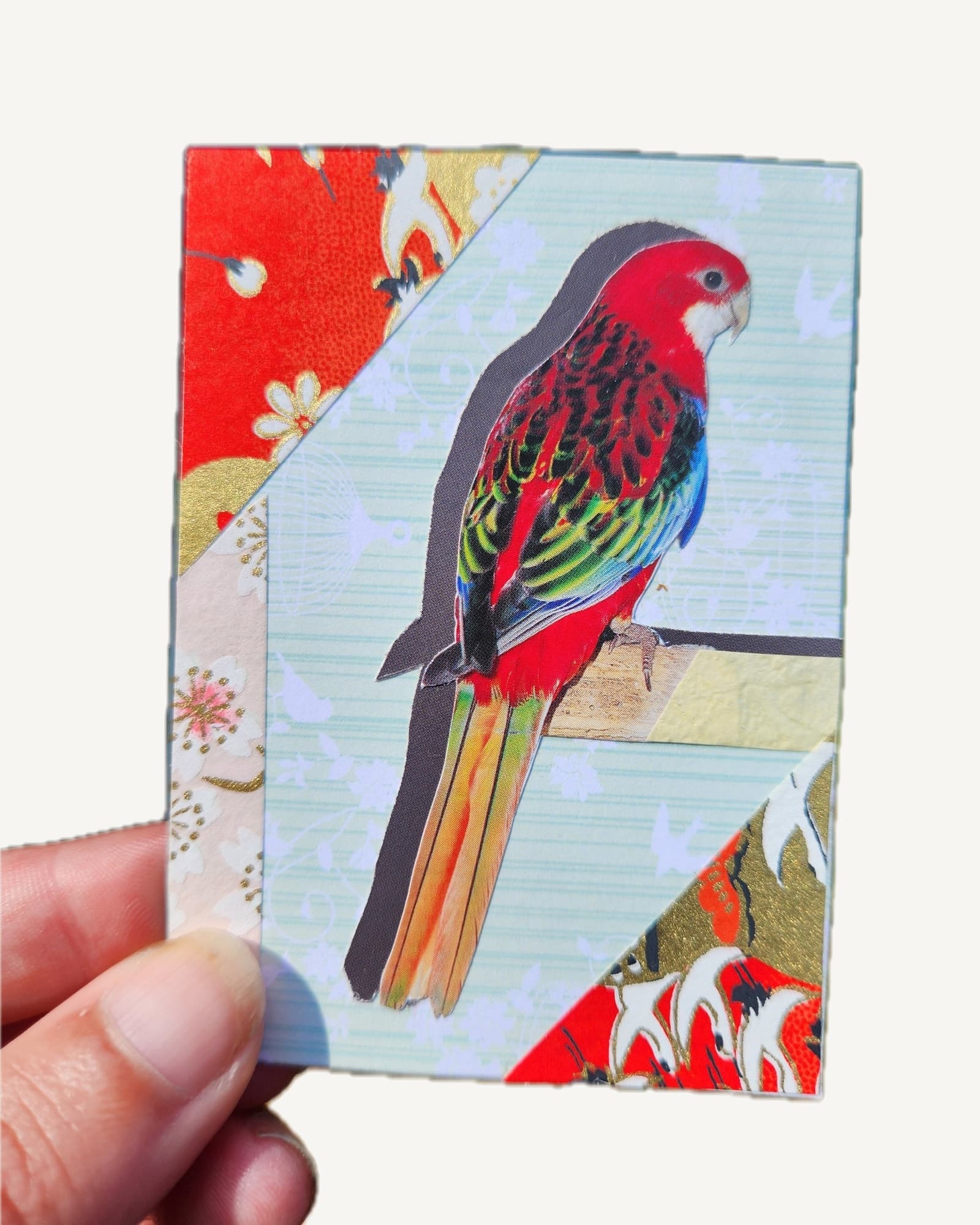 A red Eastern rosella cut by hand from a secondhand bird book, set against mint green card and red and gold origami paper.