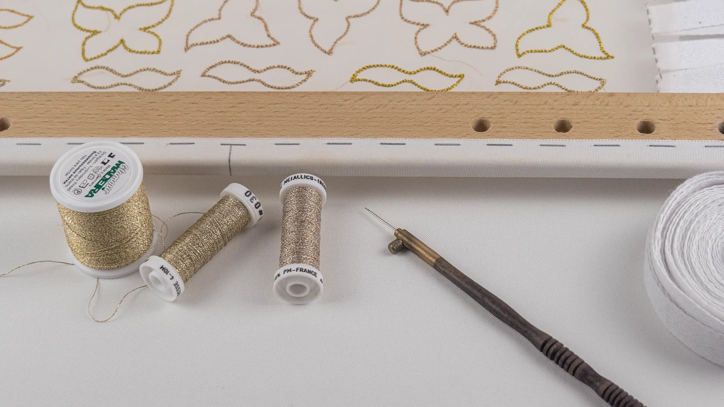Embroidery materials laid flat: gold metallic thread spools, white thread, wooden frame with perforated edge