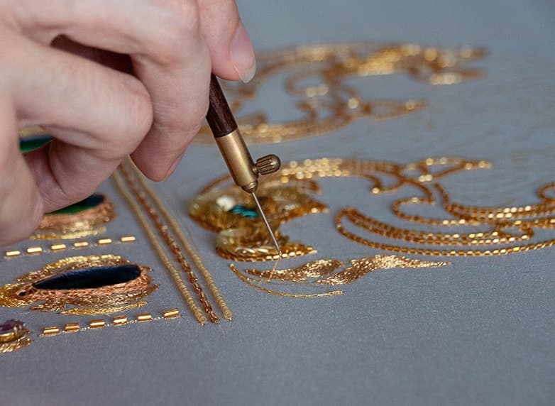 Close-up of hands performing gold thread embroidery with a fine needle on grey fabric