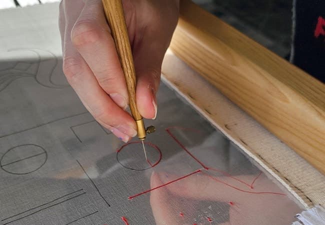 Hand holding wooden tambour hook, embroidering red thread design onto pale fabric stretched on frame.