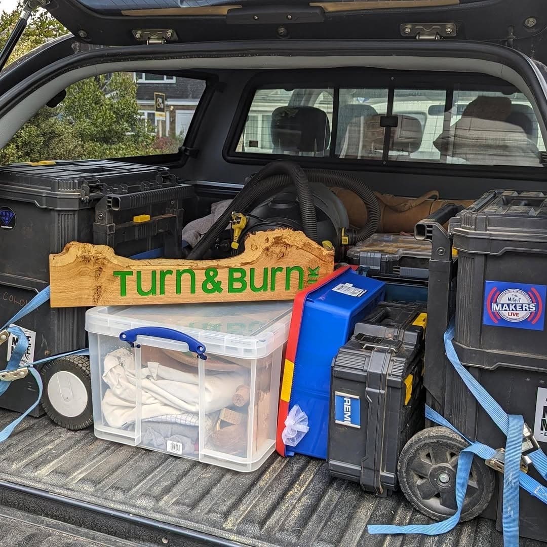 The open back of a pickup loaded with tool cases and a carved wooden Turn and Burn sign.