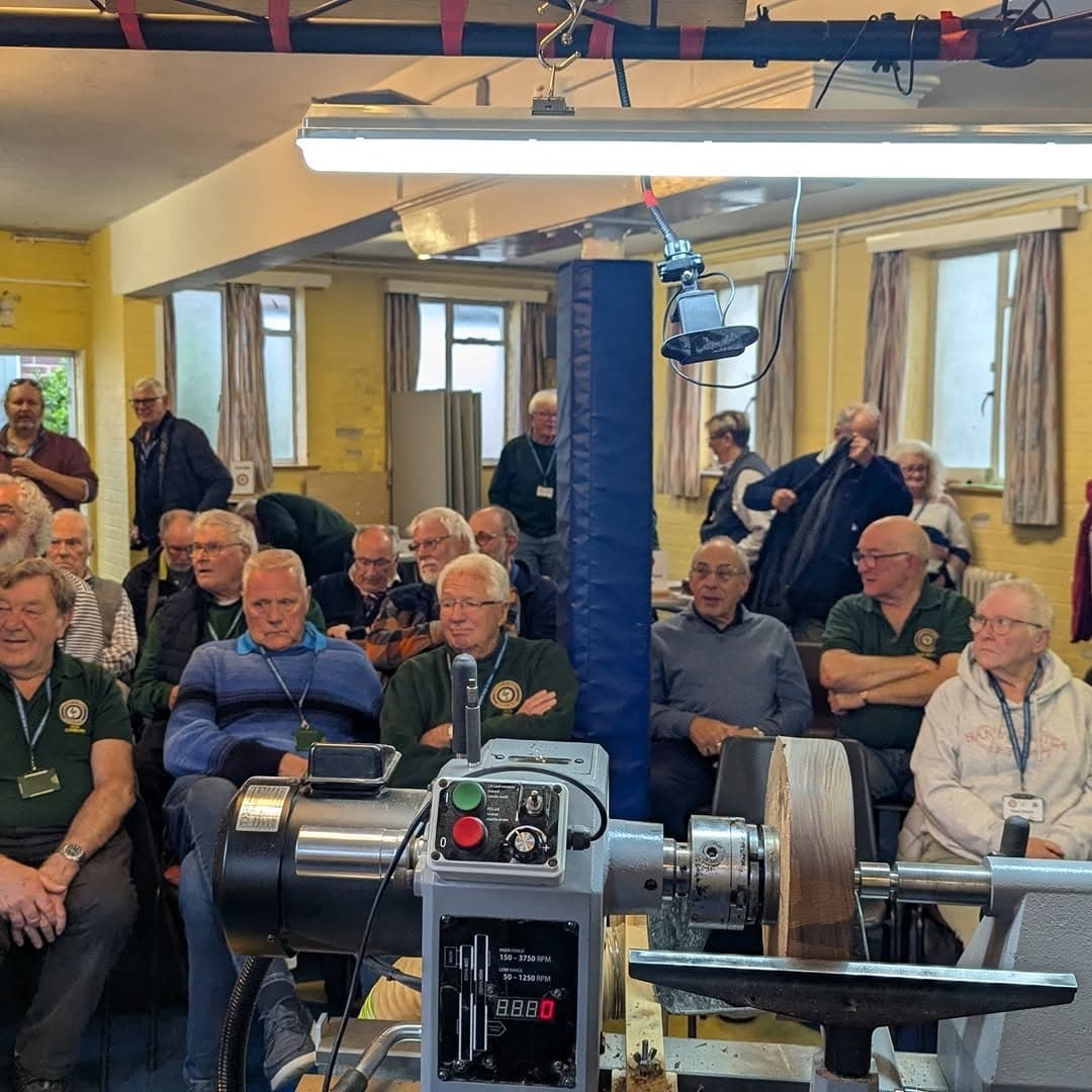 A woodturning club audience seated in rows facing a lathe set up at the front of a hall.