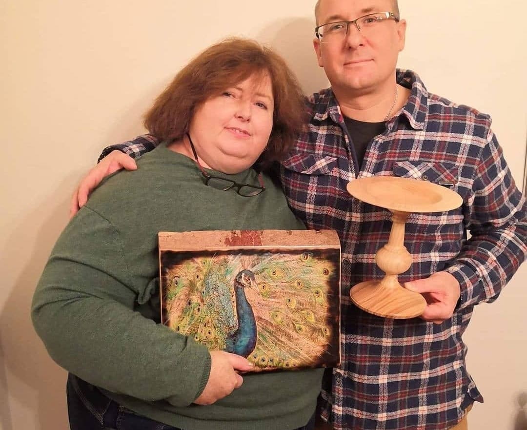Andy and Bee Matthews side by side, she holding a pyrographed peacock panel and he a turned wooden stand.