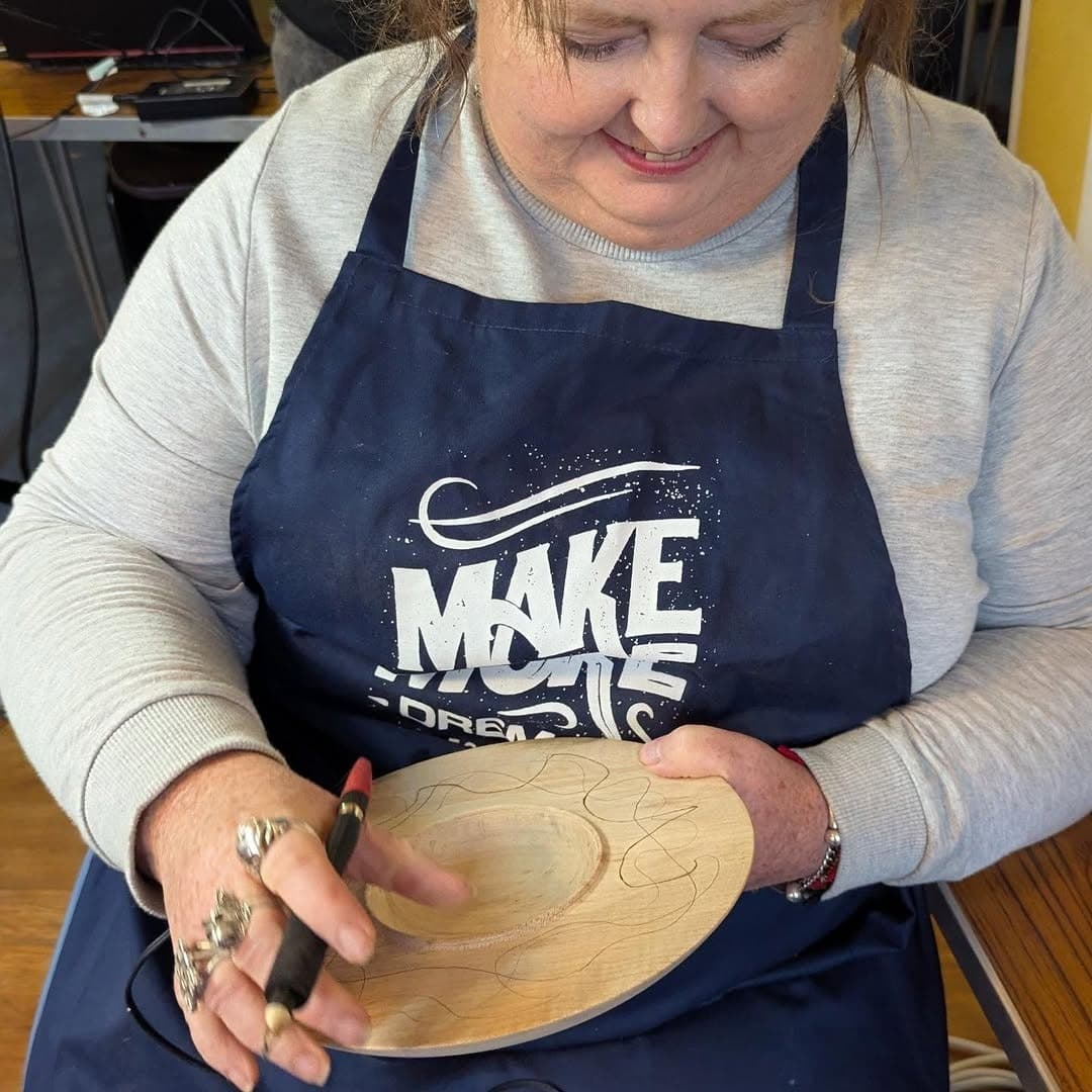 Bee Matthews holding a turned wooden plate on her lap and working a burned line into it with a pyrography pen.