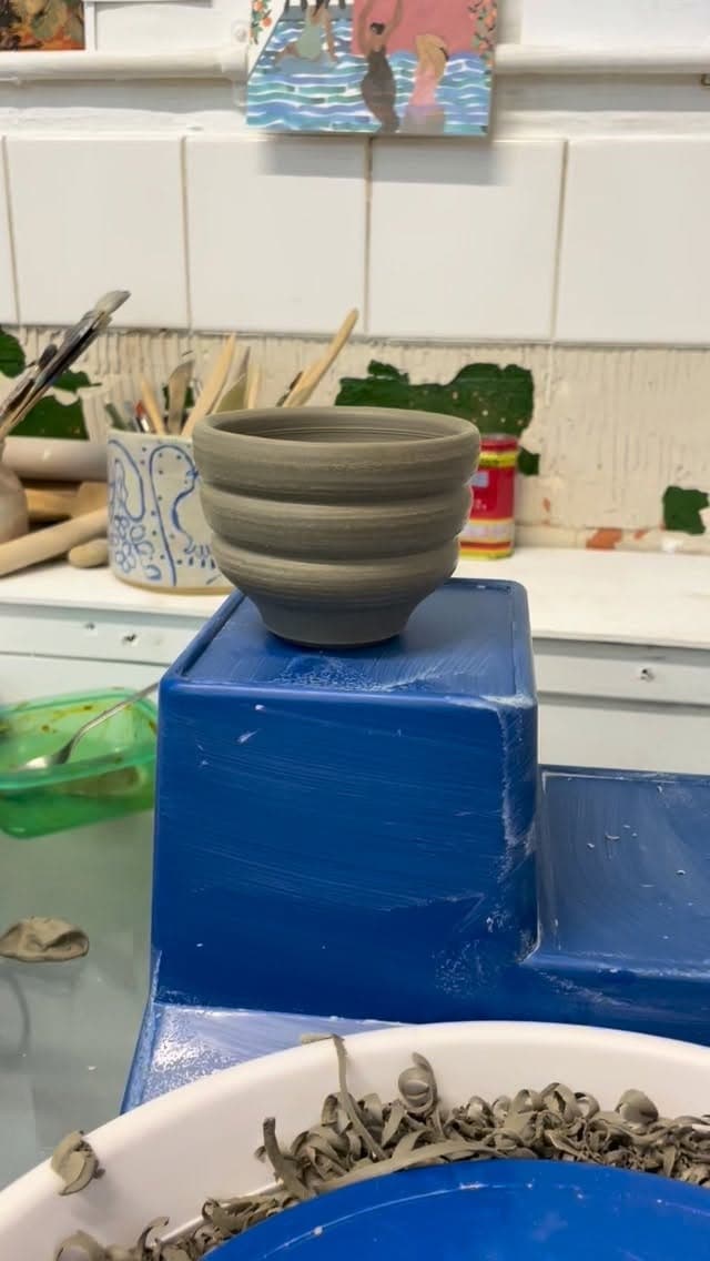 Grey ceramic bowl with horizontal grooves, displayed on blue cube in artist's studio workspace surrounded by tools and