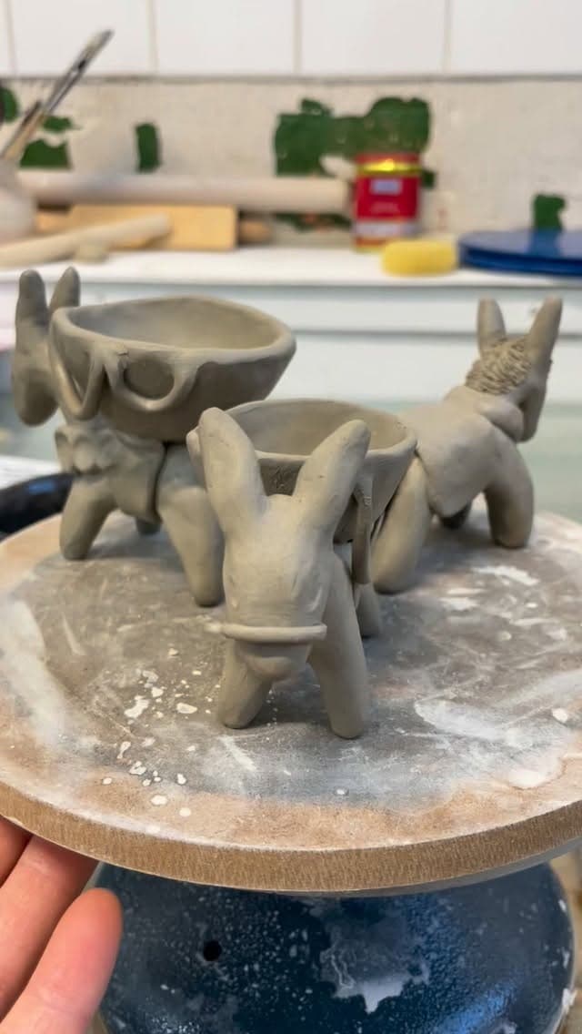 Three unfired ceramic animal figurines on wooden base: dog-shaped bowl with handles on left, rhinoceros head in center