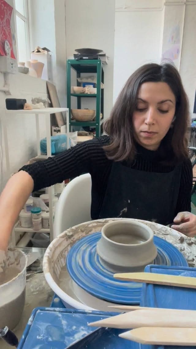 Potter at wheel working with cream-coloured clay bowl; blue-painted ceramic plate and wooden tools visible on work surface