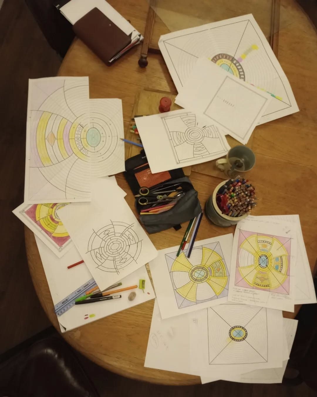Hand-drawn design cartoons for circular stained glass panels spread across a table with pencils and tools.