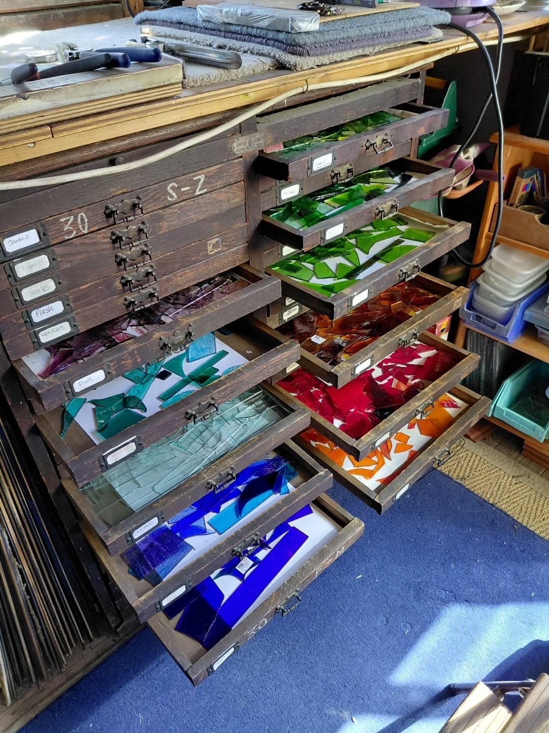 Shallow wooden drawers pulled open to show sheets of coloured glass sorted by colour.