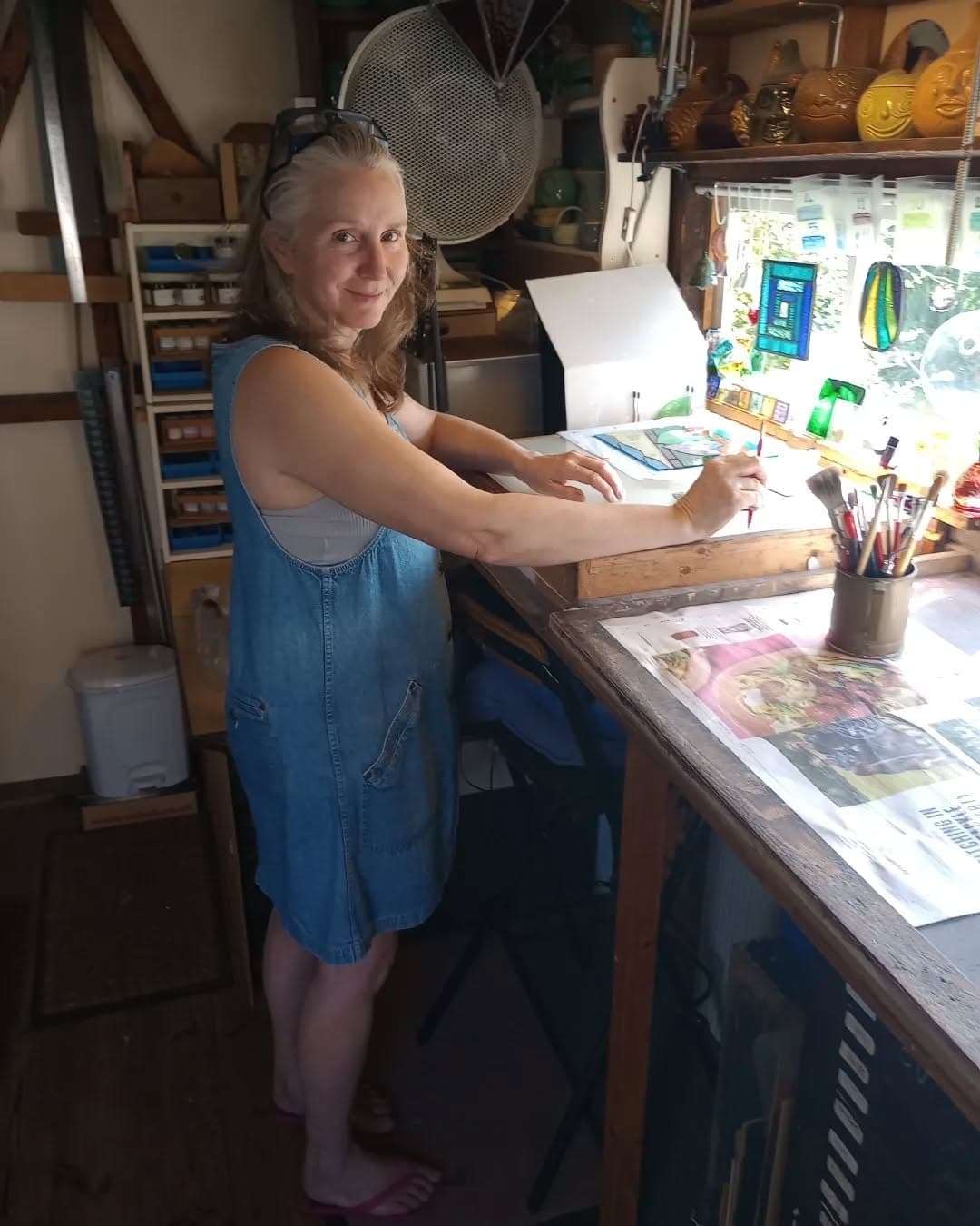 Mary Corkery standing at her workbench in her garden workshop, working on a stained glass panel by the window.