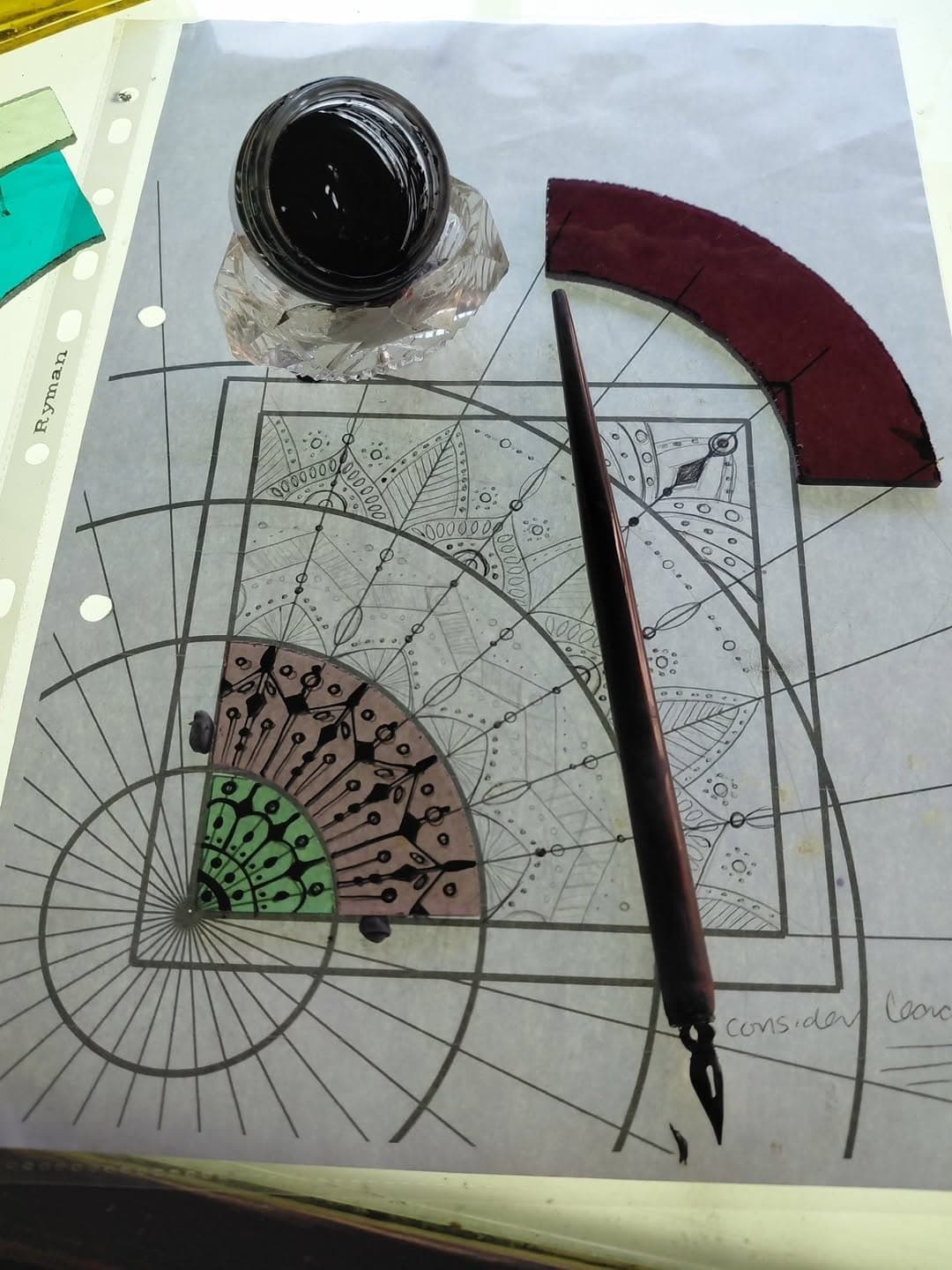 A dip pen and a pot of black glass paint resting on a paper cartoon, beside a painted fan-shaped piece of glass.