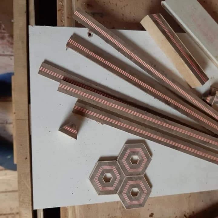 Cut strips of laminated wood with fine red lines, and three finished hexagons, on the workshop bench.