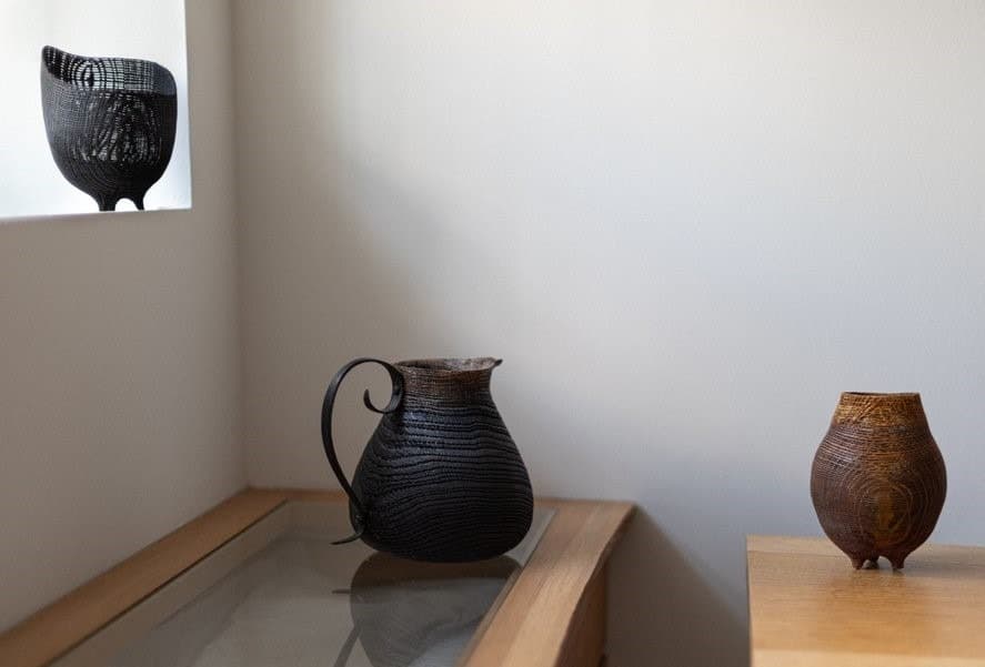 Three turned wooden vessels displayed on wooden shelves: black textured pitcher with handle, dark wood bulbous form
