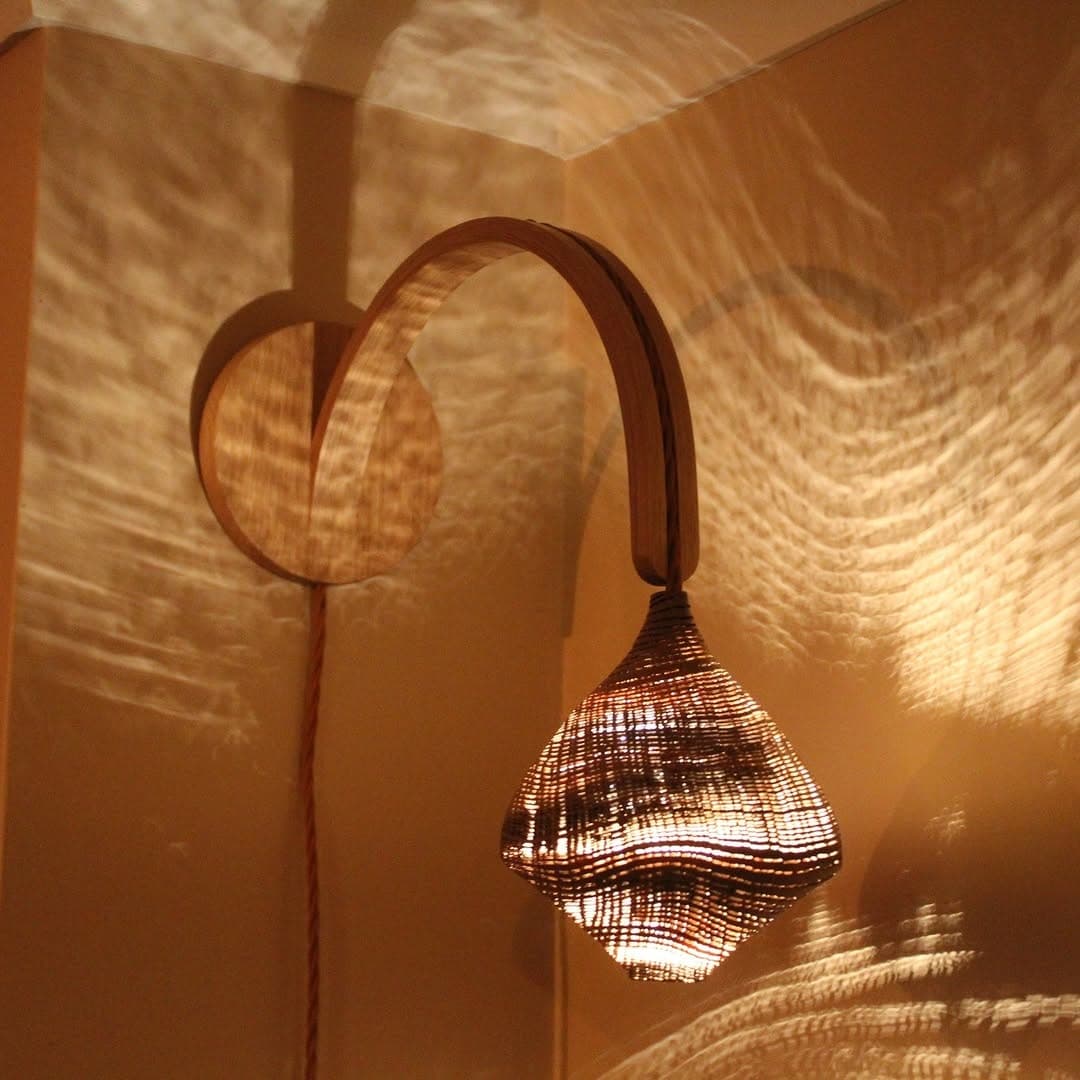 A wall lamp with a steam-bent arm and a sandblasted lace oak shade, throwing the grain across the wall