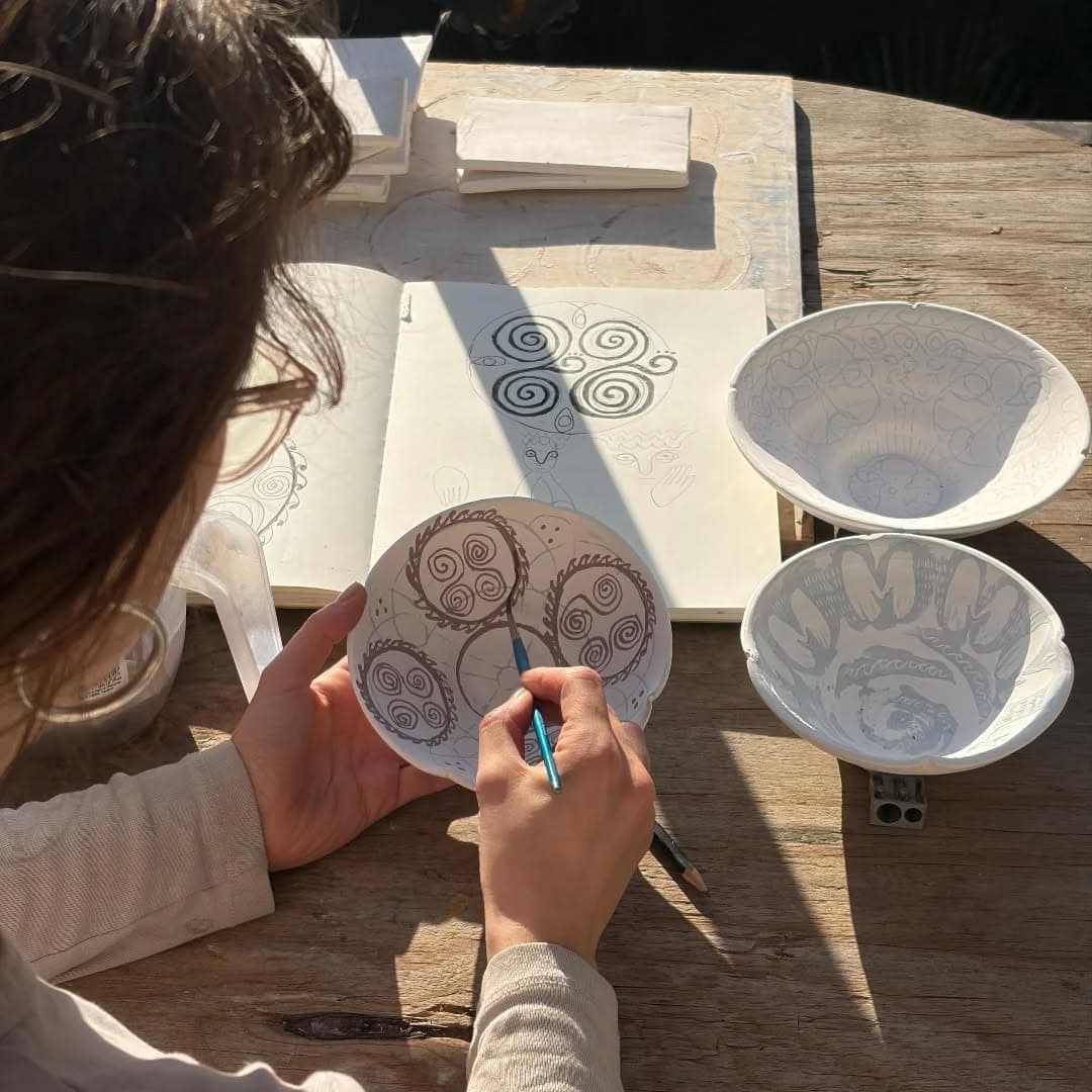 Hands decorating a ceramic disc at an outdoor bench beside an open sketchbook and two bowls