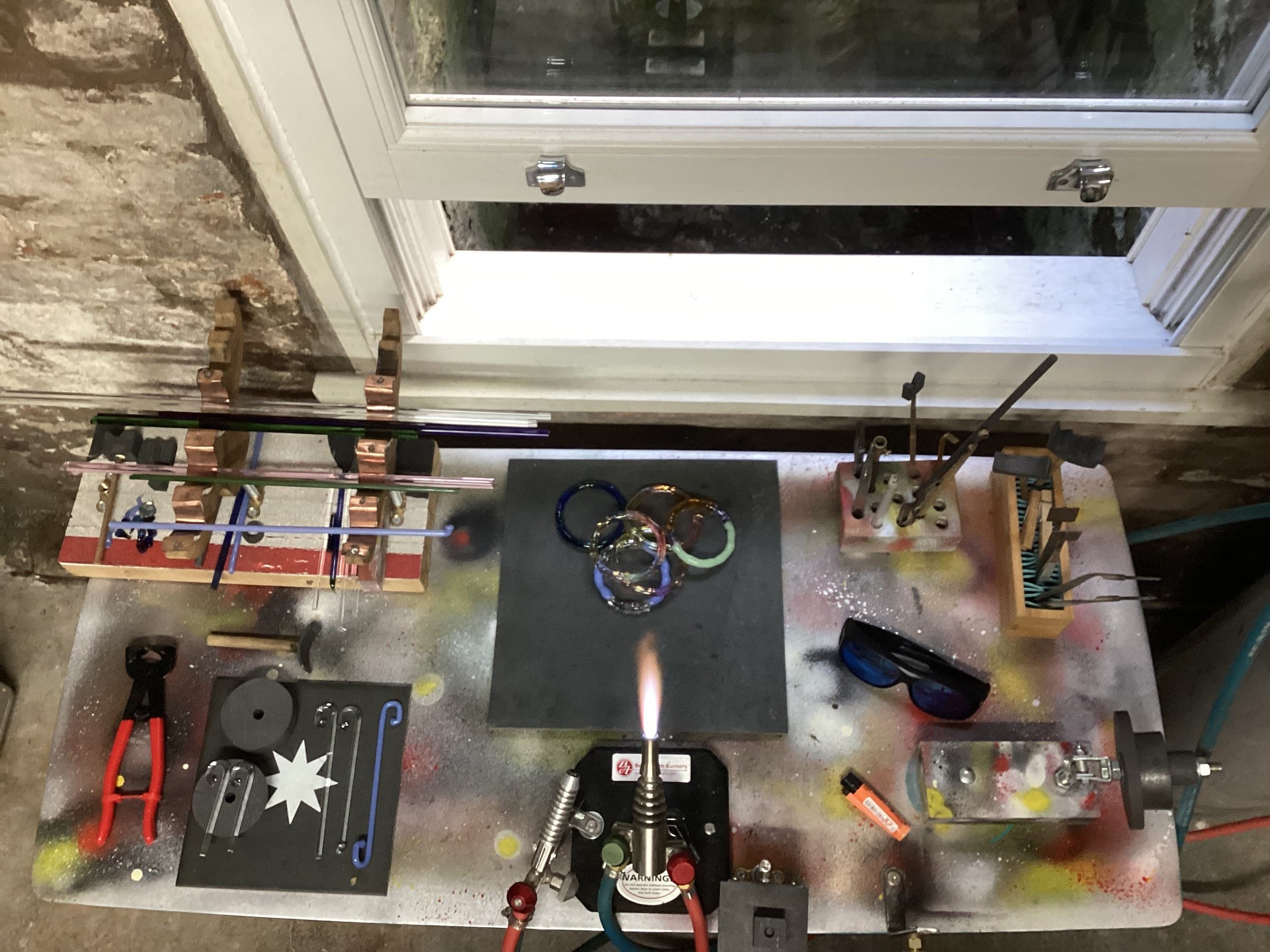 Greig Alderman's lampworking bench seen from above, with glass rods, tools and a stack of glass bangles by the window