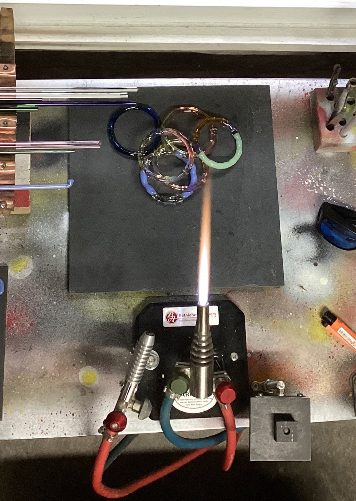 Greig Alderman's lampworking torch alight on his studio bench, with finished glass bangles on the board behind it