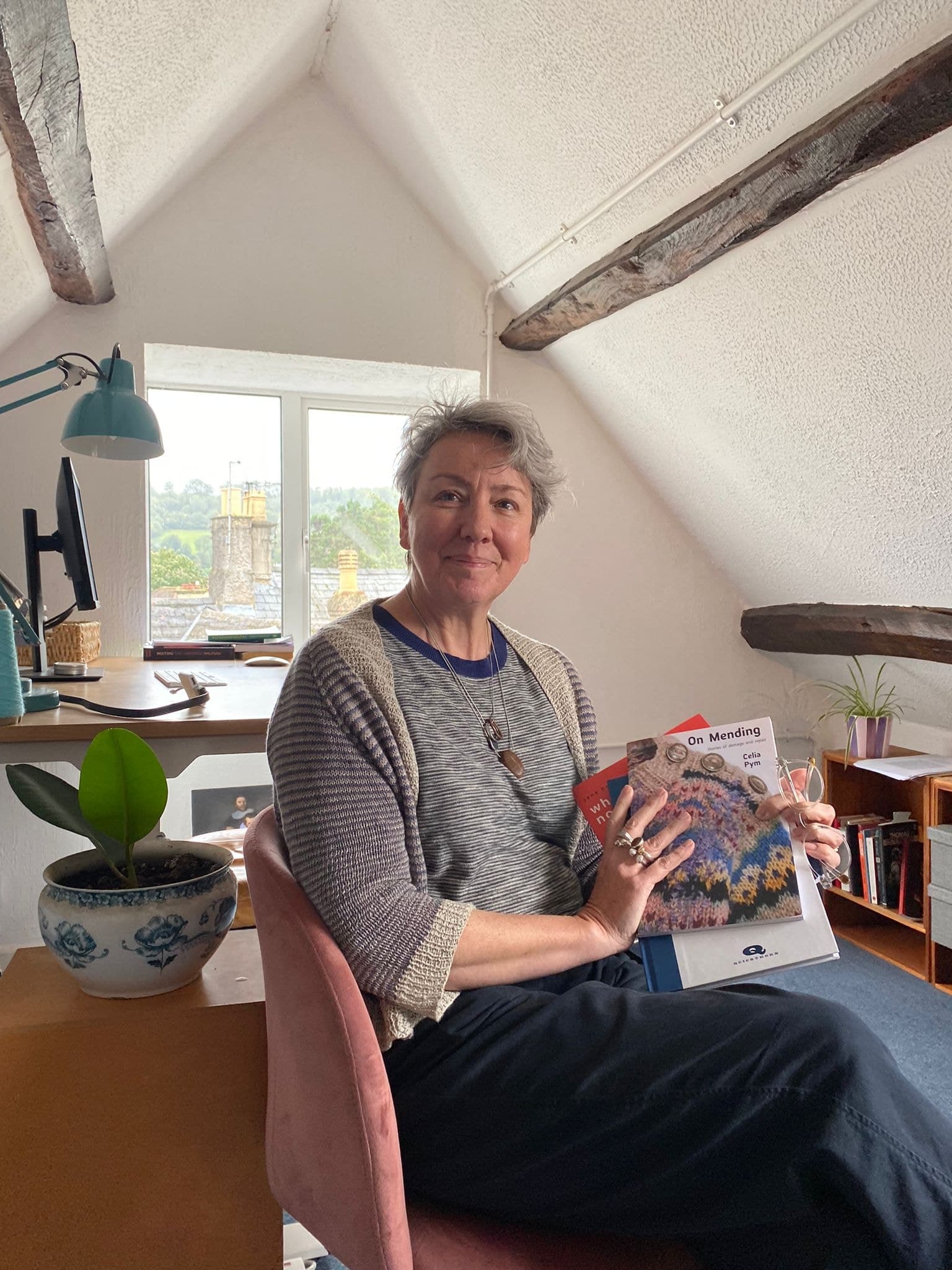 Woman holding open a book titled "On Mending" in a bright attic room with exposed wooden beams, potted plant
