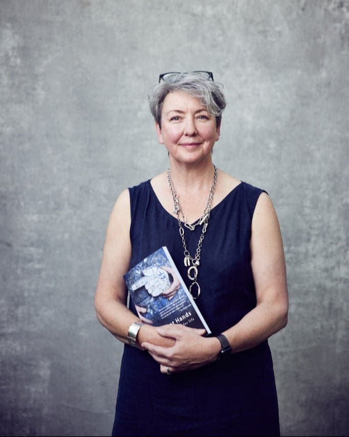 Woman with short grey hair holding a copy of the book Intelligent Hands, against a grey backdrop