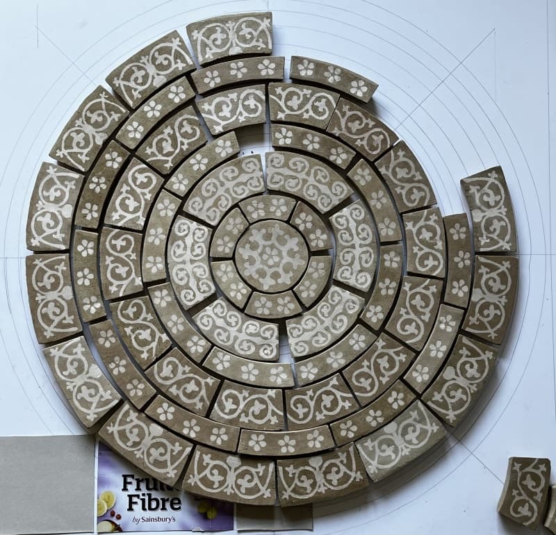 Large circular pavement of curved unfired tiles in rings of white foliage scrolls and flowers, laid on a paper template