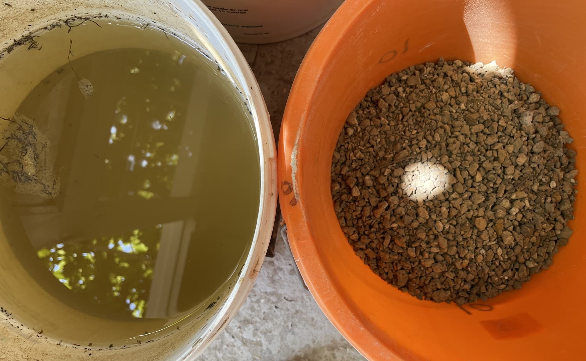 Bucket of clay slip settling in water beside an orange bucket of dried, crushed garden clay