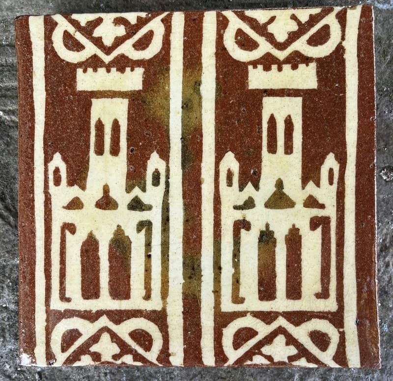Red clay floor tile inlaid in white with two castle towers under battlements, the glaze patched honey and green