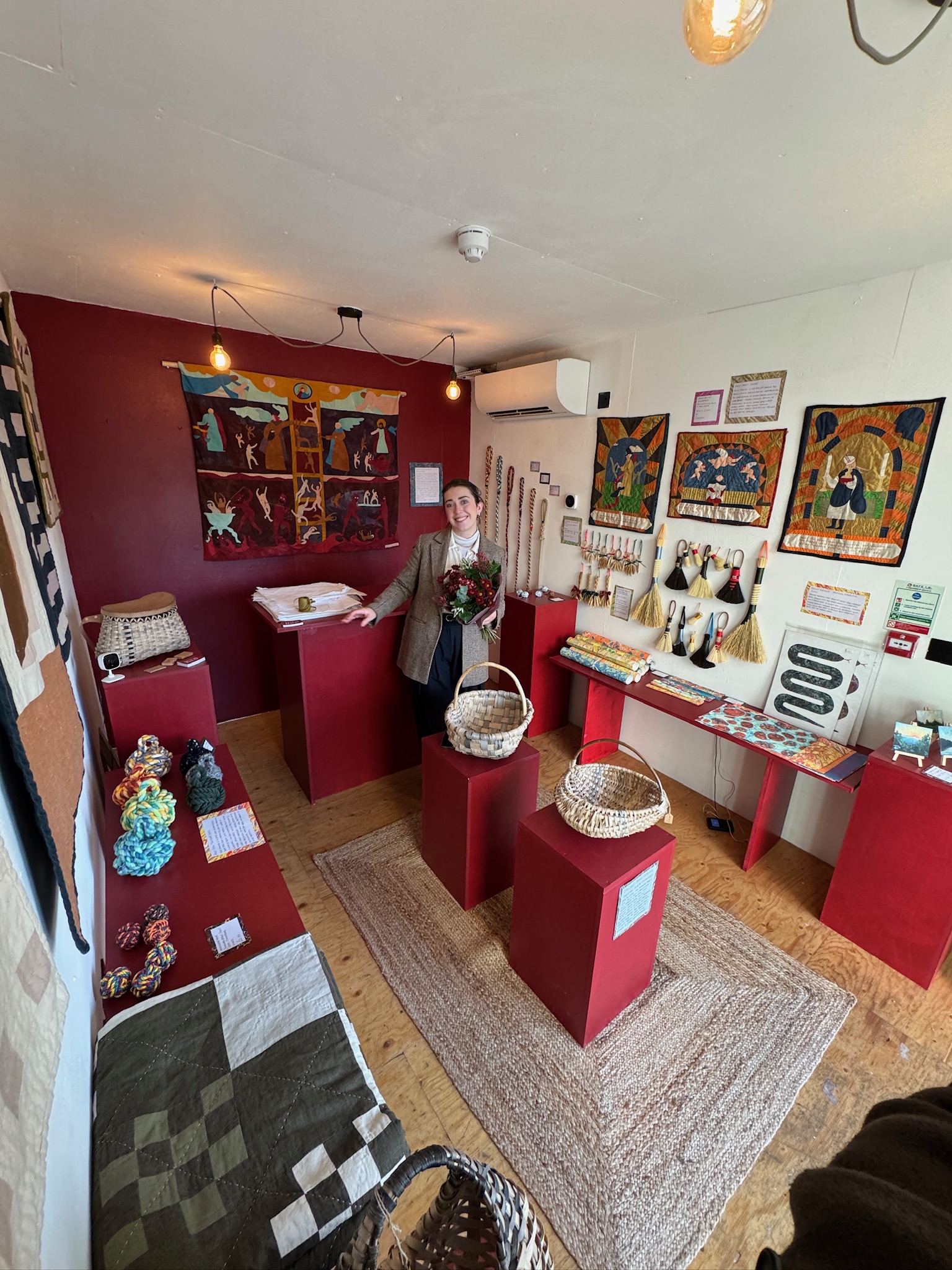 Rosie Thompson inside The Haud pop-up shop, surrounded by red plinths showing baskets, ropes, brushes and quilted hangings