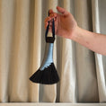 Load image into Gallery viewer, Hand holding a brush with a black bristle head and blue handle against a beige curtain background
