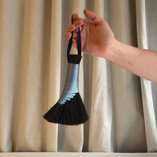 Hand holding a brush with a black bristle head and blue handle against a beige curtain background