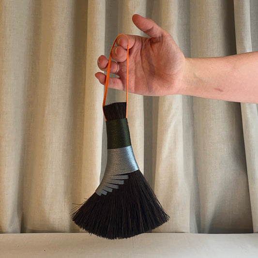 Hand holding a black and green brush with an orange handle against a beige curtain background