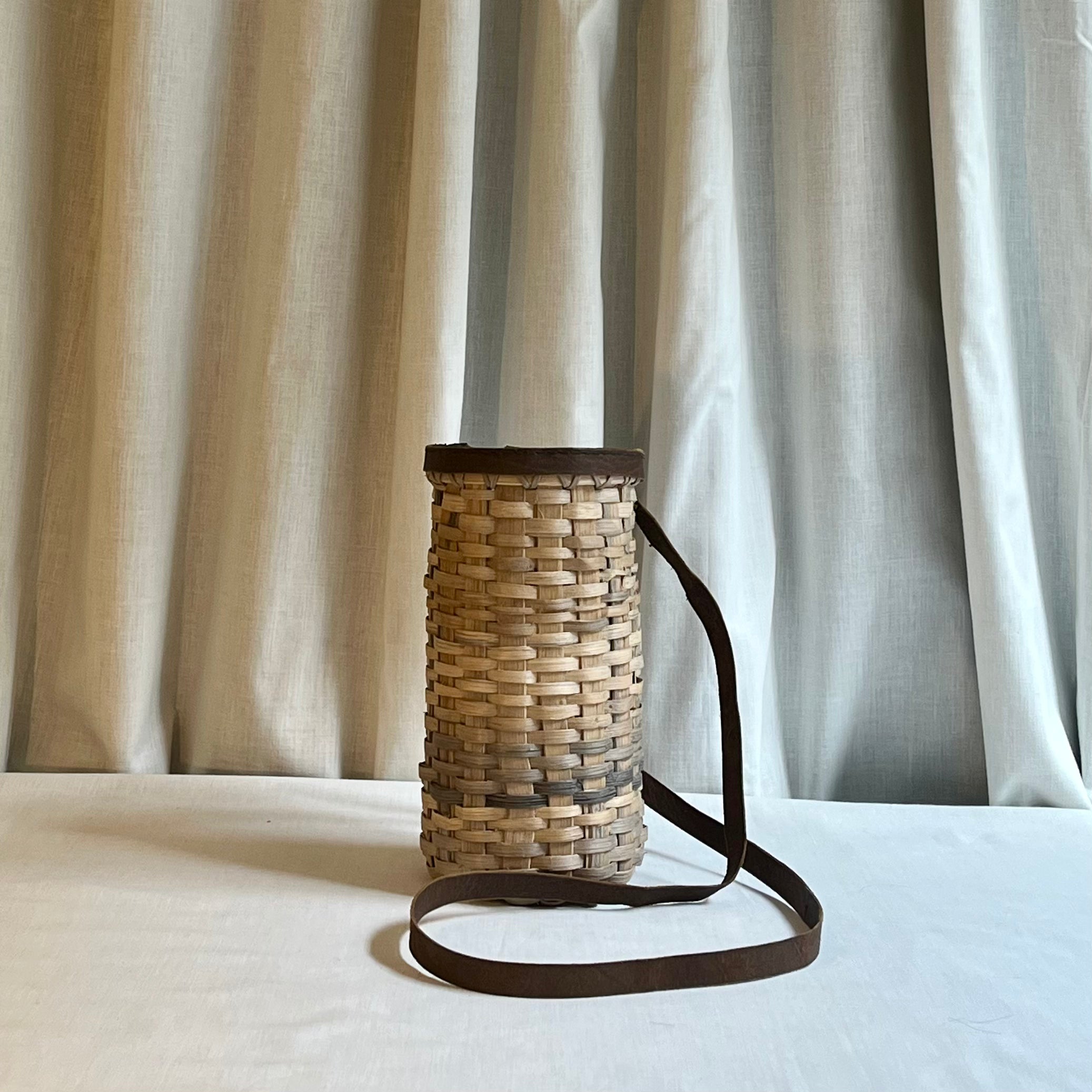 Woven basket with a strap on a white surface with curtains in the background