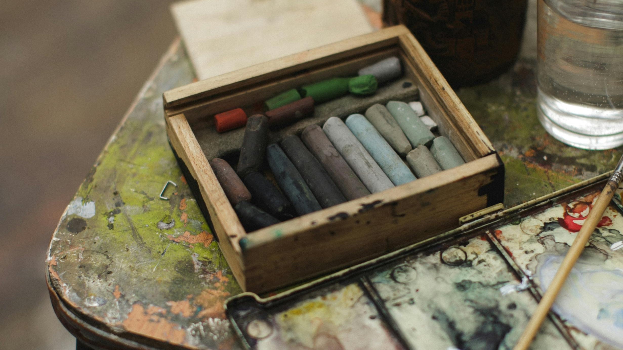 Art supplies including pastels, a palette, and a glass of water on a rustic table.