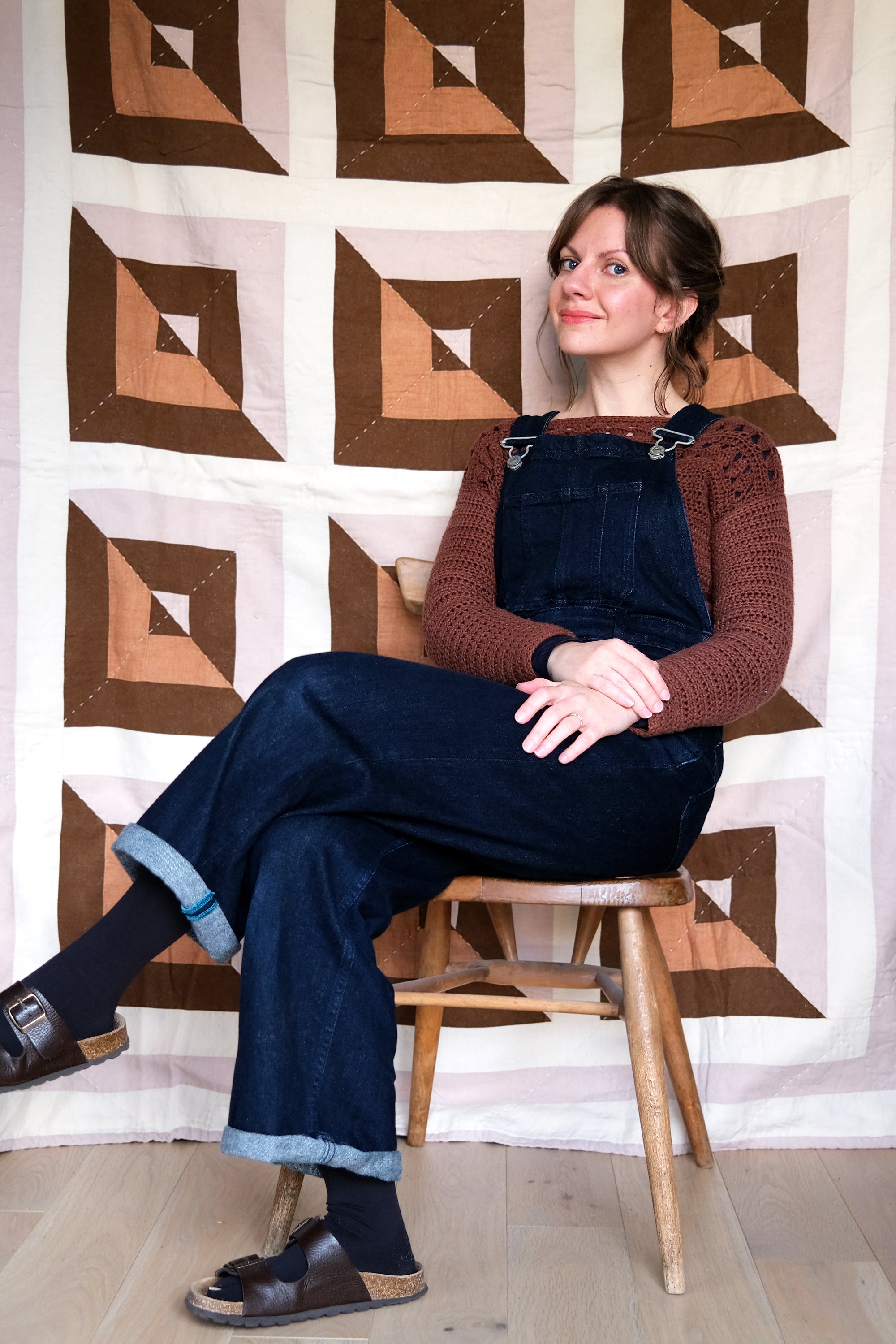 Woman sitting on a stool wearing a brown sweater and blue denim overalls against a geometric-patterned wall.