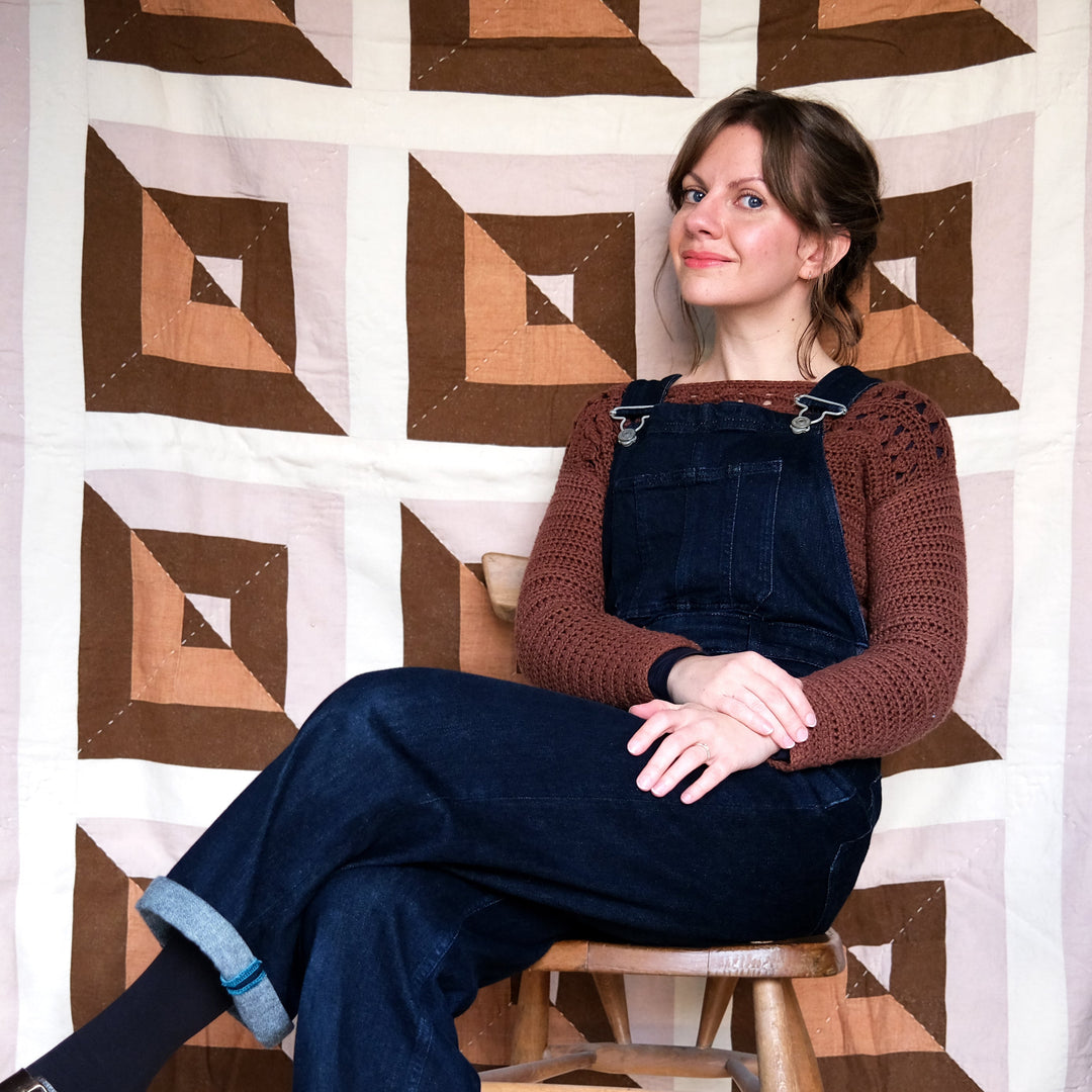 Woman sitting on a stool wearing blue overalls and a brown sweater against a geometric-patterned wall.